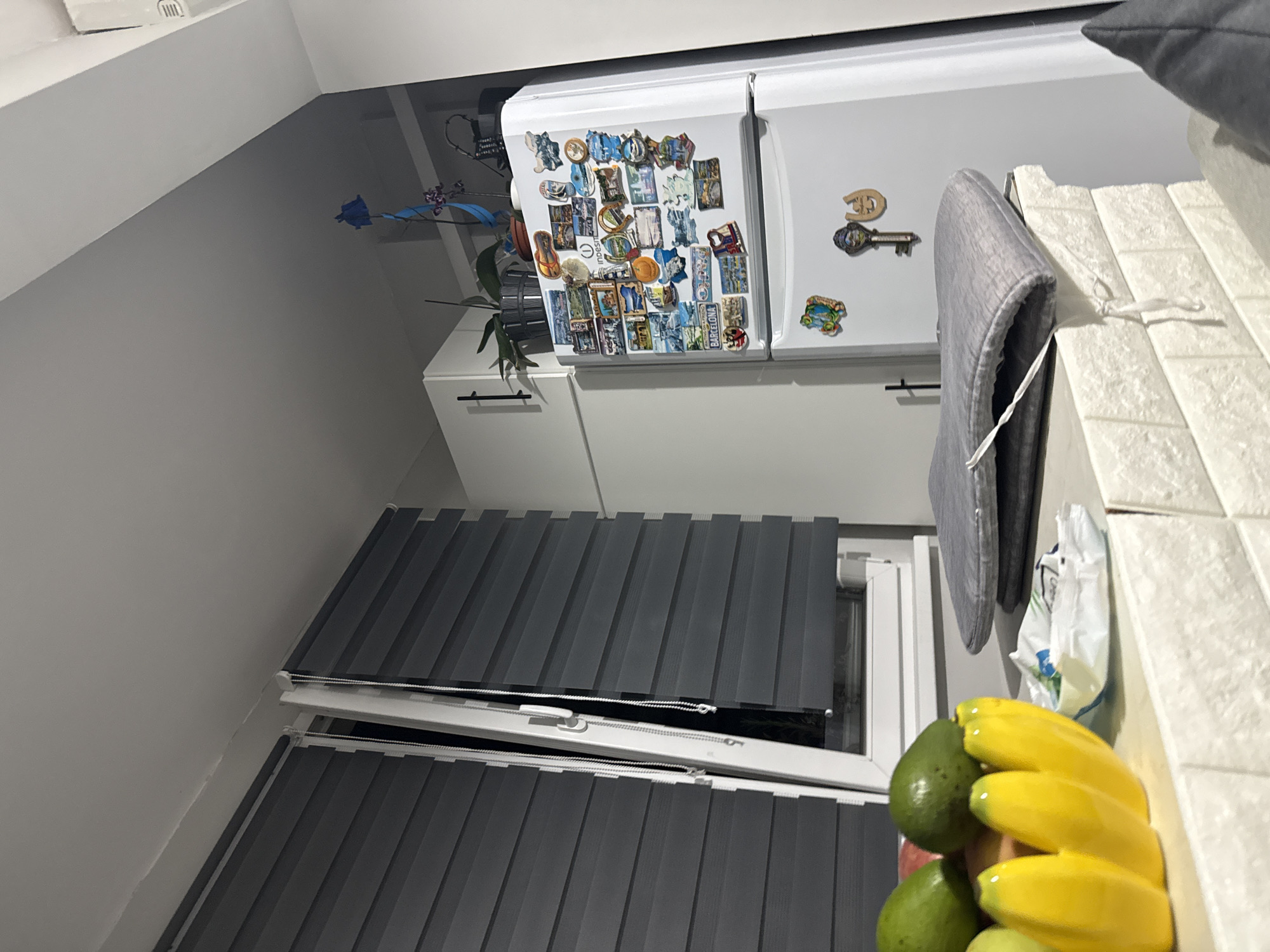 A residential interior is shown. The focal point is the kitchen area, which features a white refrigerator adorned with magnets. Next to the refrigerator, there is a white cabinet. A window is visible with gray blinds partially drawn. On the counter are a bag of bananas and avocados, and a gray coat. The location is Stara Zagora, Bulgaria.