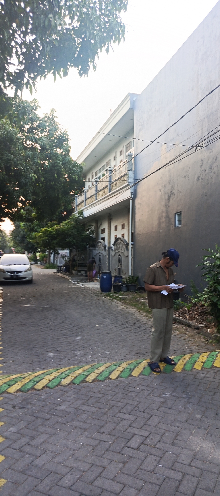 In the city of Sukaresmi, Indonesia, on a paved street with a textured brick pattern, a man wearing a brown short-sleeved shirt, beige pants, a blue baseball cap, and dark sandals stands facing left, holding a white document in his hands. The man appears to be examining the document.
To his left, a white car is parked on the street. Behind the man, a two-story building with a white facade and balconies with ornate railings is visible. A large, dark gray wall with a small square window is adjacent to the building. The street has a speed bump painted with alternating yellow and green stripes.
In the background, mature trees with dense foliage are present, and to the right, there is an area with dry leaves and vegetation. Two other individuals are partially visible in the distance near a gate. A large blue barrel is situated on the side of the road. The lighting suggests it is either early morning or late afternoon.