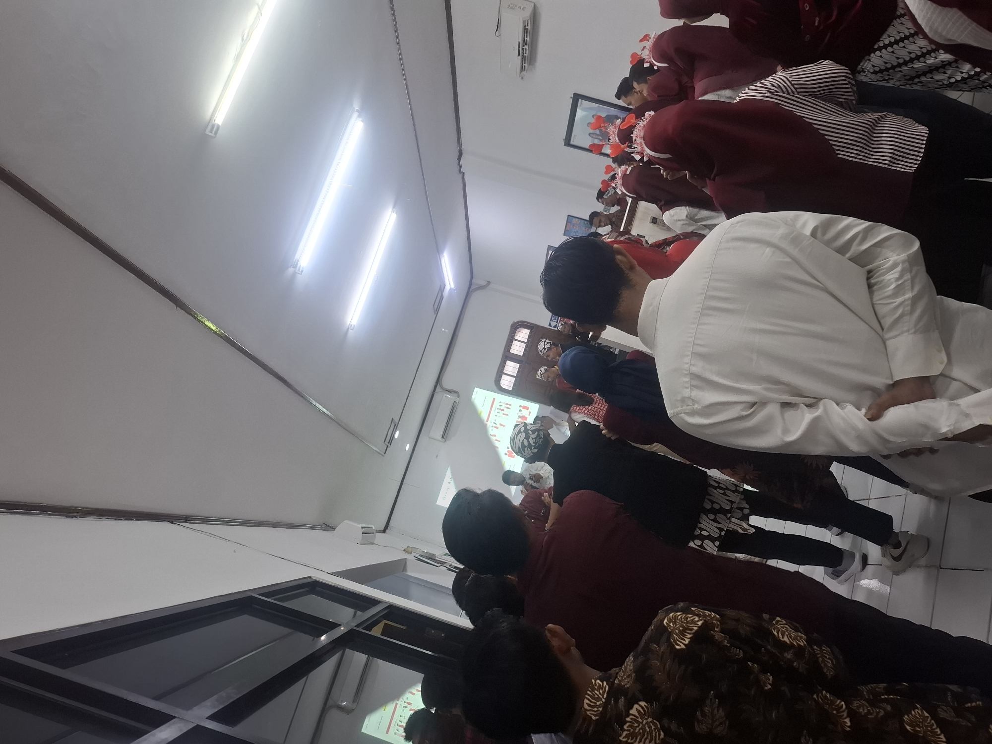 The image depicts a gathering of people indoors, likely in a classroom or meeting room. The room is brightly lit by fluorescent lights on the ceiling. A projector screen is visible on the wall, displaying a presentation with graphics and text. Several individuals are facing the screen, some wearing traditional Indonesian attire, including batik patterns and headwear. Others are dressed in more casual clothing, with some wearing maroon or dark-colored shirts and headscarves. The overall atmosphere suggests an educational event or a cultural celebration. While the specific location within Lamongan, Indonesia, cannot be determined from the image alone, the presence of Indonesian attire and the context of a gathering point to a local event.
