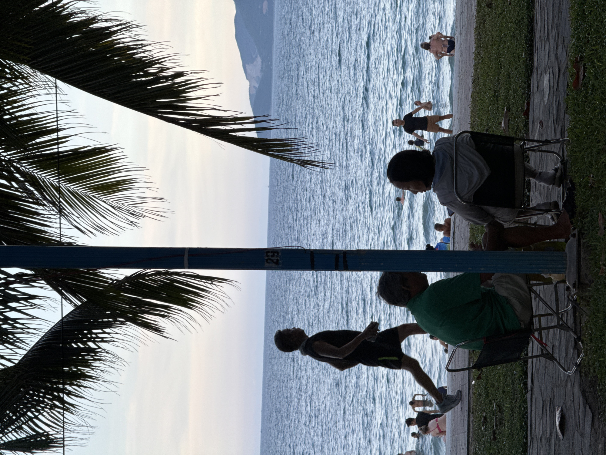 The image depicts a scene at a beach in Nha Trang Ward, Vietnam, likely during late afternoon or early evening given the soft light. In the foreground, silhouetted against the water, are two individuals seated on folding chairs, facing away from the viewer towards the sea. One person wears a green shirt and khaki shorts, while the other is in a grey hoodie. A blue pole stands between them, with a number '29' visible on it.
Beyond the seated figures, a man in dark athletic wear and shorts is captured in mid-stride, running along the paved walkway that borders the beach. The ocean stretches out behind him, with a gentle swell and numerous people in the water, some swimming or wading. In the distance, a hazy coastline with hills is visible. Palm fronds hang from the upper portion of the frame, casting shadows and framing the view of the beach and sea. The overall atmosphere is relaxed and suggests a leisure activity.