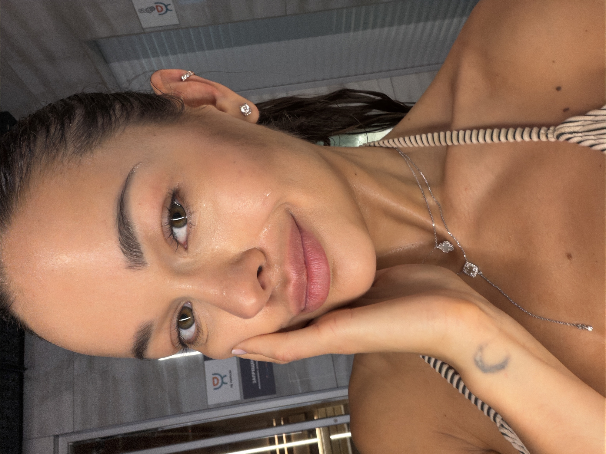 A close-up selfie shows a woman with wet, dark hair pulled back, appearing dewy or perspiring. She has brown eyes and is looking directly at the camera with a slight smile. Her left hand gently touches her left cheek. She wears a striped top, possibly a swimsuit, with a coiled strap visible on her right shoulder. Jewelry includes a diamond-like stud and a cuff earring on her right ear, and two delicate silver necklaces with a clover charm and a square pendant. A faded crescent moon tattoo is visible on her left inner wrist. The background features light gray tiled or paneled walls with vertical strip lighting reflections. Two signs in Russian are visible: a red and white logo resembling the "World Class" fitness club brand with the text "НЕ КУРИТЬ" (NO SMOKING) above a dark gray sign displaying "ЗАПРЕЩЕНО" (FORBIDDEN). This indicates the location is likely within a World Class fitness club or spa in Moscow, Russia.