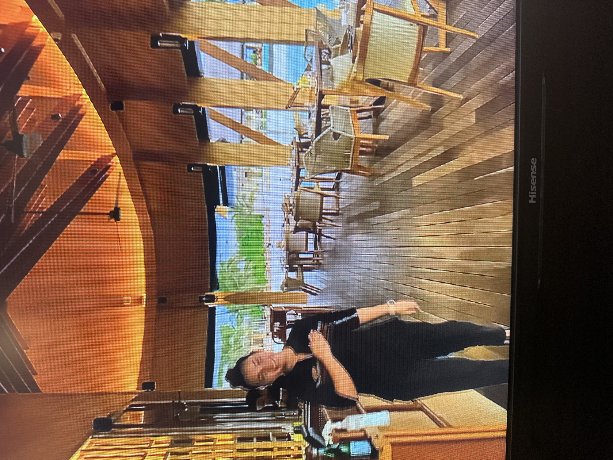 The image shows a woman standing on a wooden floor in what appears to be an open-air restaurant or dining area. She is wearing a black uniform with a white logo on the left breast and black pants. She has her left hand placed over her chest and is smiling at the camera. Behind her, tables and chairs are set up, and beyond that, there is a view of palm trees and water, suggesting a tropical or coastal location. The architecture of the building features exposed wooden beams and a warm, orange-toned ceiling. A bottle and a dispenser are visible on a counter to the left of the woman. The image is taken from a slightly low angle, and the Hisense logo is visible at the bottom of the frame. The scene is not located in Kyiv, Ukraine.