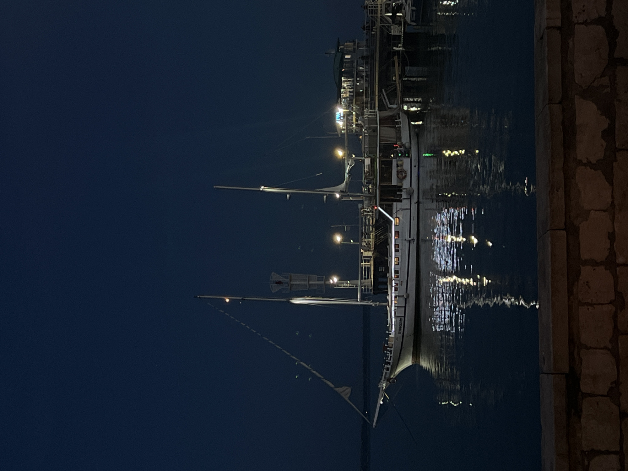 A white boat with multiple decks is docked at night. The boat is illuminated by interior lights and string lights, and its reflection shimmers on the dark water. A pier with structures and lights is connected to the boat. The sky is a deep, dark blue, indicating nighttime. In the foreground, a stone wall made of rough-hewn rectangular blocks runs horizontally across the bottom of the frame. The scene is set in a harbor or waterfront area.
