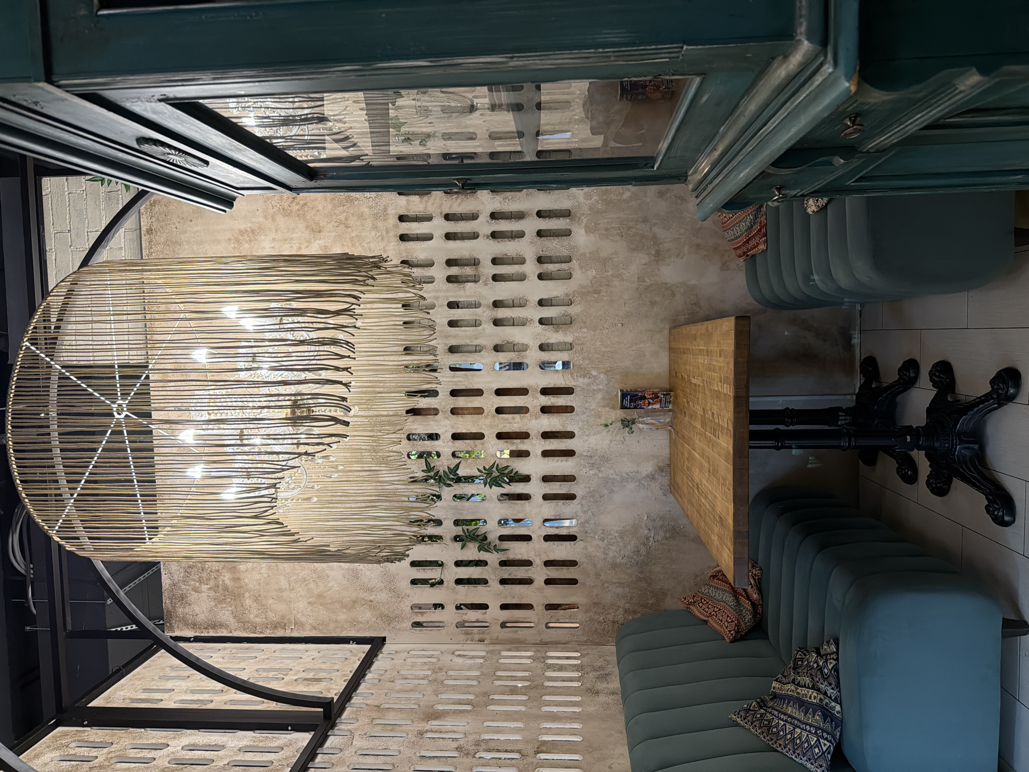 This image displays an interior view of a commercial space, likely a cafe or restaurant, captured in a portrait orientation that has been rotated 90 degrees clockwise. The setting combines rustic, industrial, and bohemian design elements, creating an inviting ambiance.
The scene is dominated by a textured, light-colored wall with a distressed, concrete-like finish. This wall features a repeating pattern of horizontal, rectangular cutouts, reminiscent of decorative breeze blocks. Small clusters of green plants subtly emerge from some of these openings, adding a natural touch.
To the right, two deep teal or dark green upholstered booths or bench seats are arranged along a wall, facing a wooden tabletop supported by a heavy, ornate black cast-iron base. Decorative throw pillows with intricate, patterned designs (one reddish-brown, one blue and white) are placed on the seats, enhancing the cozy feel.
Above the booths and table, a dark teal-colored wooden structure, possibly a cabinet or shelving unit, is mounted. It features glass panels with reflections of the interior, and its painted surface shows some wear and decorative molding.
To the left, a large, circular pendant light fixture hangs from the ceiling. It is crafted from woven natural material, resembling wicker or rope, and houses multiple exposed light bulbs that emit a warm glow. Beneath this fixture, a black metal structure, possibly a frame or part of a partition, features a distinct curved element. The floor throughout the visible area is a light-colored, possibly concrete or tiled, surface that incorporates patterns similar to the cutouts on the main wall.
No people are visible in the image, suggesting the space is currently empty or awaiting customers. The artificial lighting from the fixture contributes to a warm and intimate atmosphere. A small item on the wall above the rearmost booth has text that includes "CHAI." The time of day is not discernible due to the indoor setting and artificial illumination. The location is an unknown indoor commercial establishment.