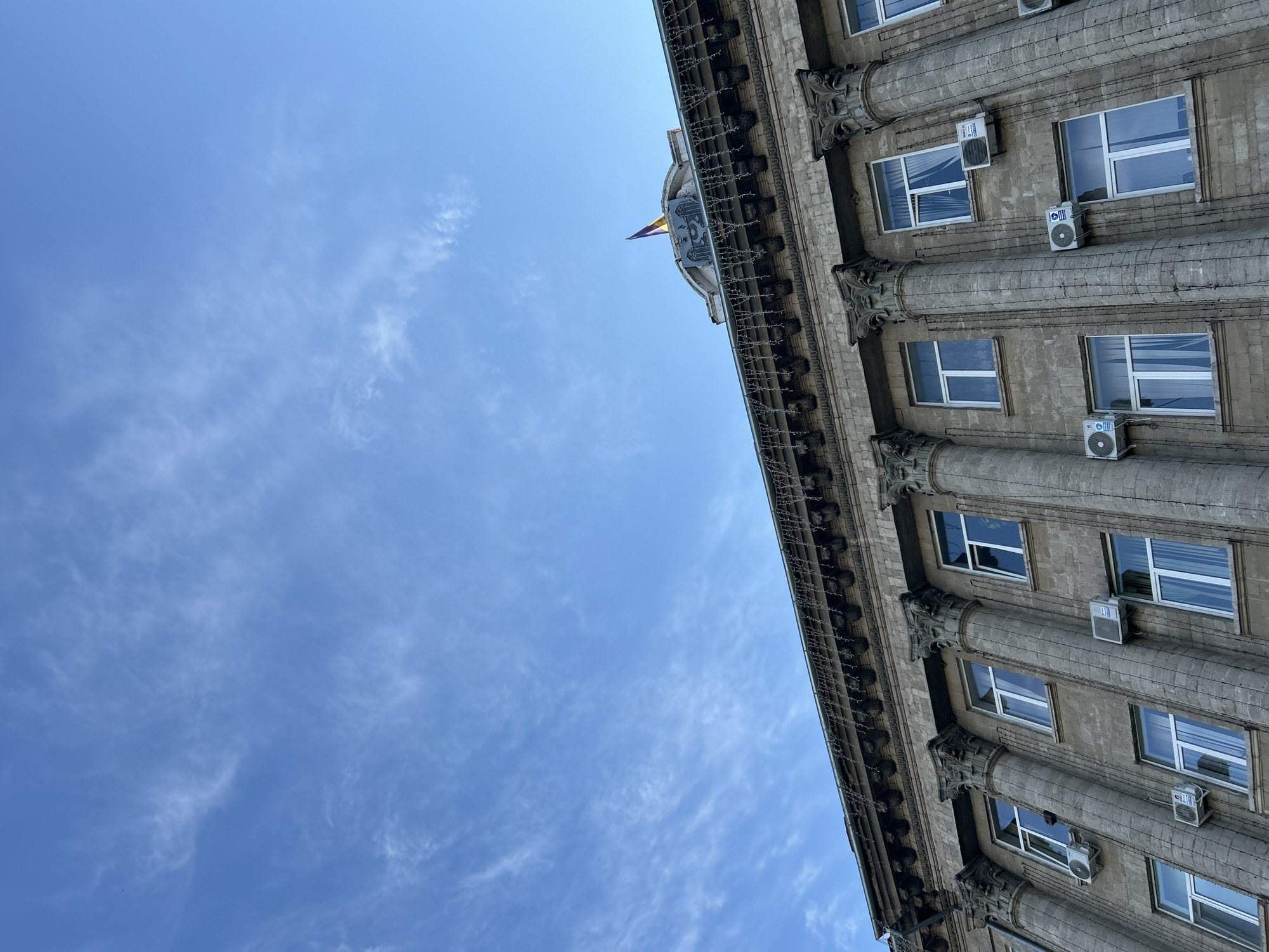 The image shows a close-up, low-angle view of the exterior of a large, old, stone building. The architecture features classical elements such as large columns with ornate capitals running vertically along the facade. The building has multiple floors with rows of identical windows, each with white frames and light-colored curtains visible. Several outdoor air conditioning units are mounted on the exterior wall between the windows. The roofline is visible at the top, adorned with decorative molding and what appear to be strings of festive lights, possibly for a holiday. At the very top center of the building, on a curved section of the roof, a flag is visible, mounted on a flagpole. The flag has horizontal stripes of yellow and purple and is partially obscured by a decorative architectural element. The sky above is a clear, bright blue with wispy, white cirrus clouds scattered across it. The scene appears to be set in the city of Bălți, Moldova, likely a public or administrative building given its imposing architecture and the presence of a national or regional flag. There are no people visible in the frame.