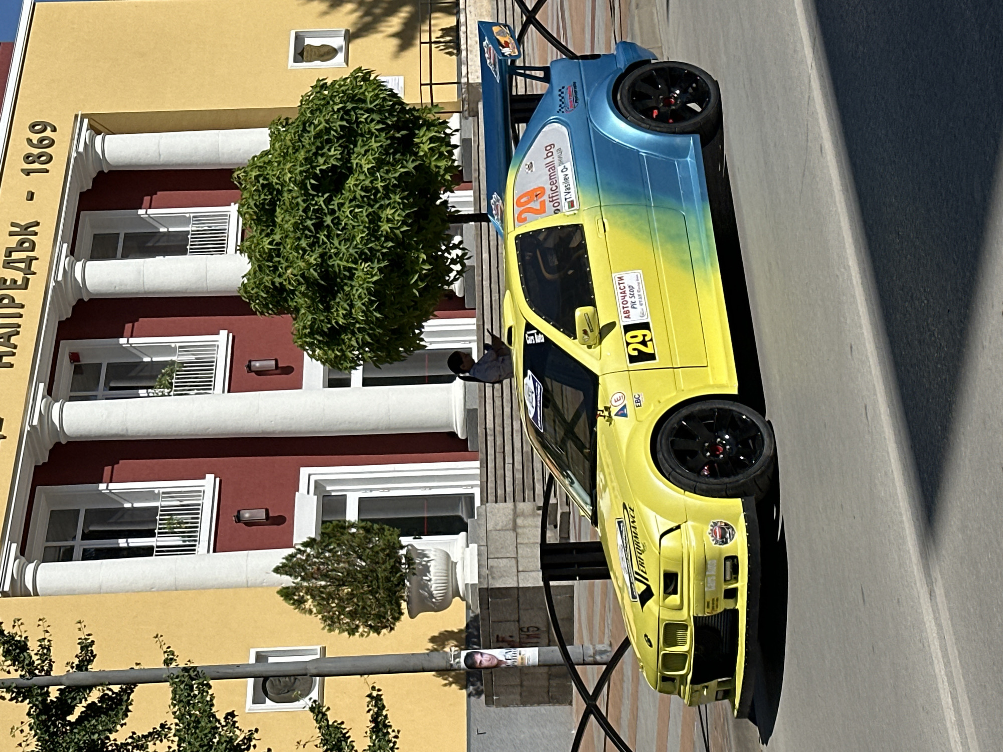 This is a daytime, eye-level, medium shot of a modified yellow and blue BMW M3 race car parked on a street in front of a yellow and red building in Gorna Oryahovitsa, Bulgaria. The building has classical architectural elements, including large white columns and ornate trim. The car has racing decals and sponsor stickers, including the number 29 prominently displayed. It features a large rear spoiler and wide body kit. The street in the foreground is asphalt with white lane markings. Behind the car, a black metal fence separates the street from a stone-paved area with a tree in a planter and stairs leading up to the building. A person is seated on the stairs, looking at a device. A light pole with a poster is visible to the left.