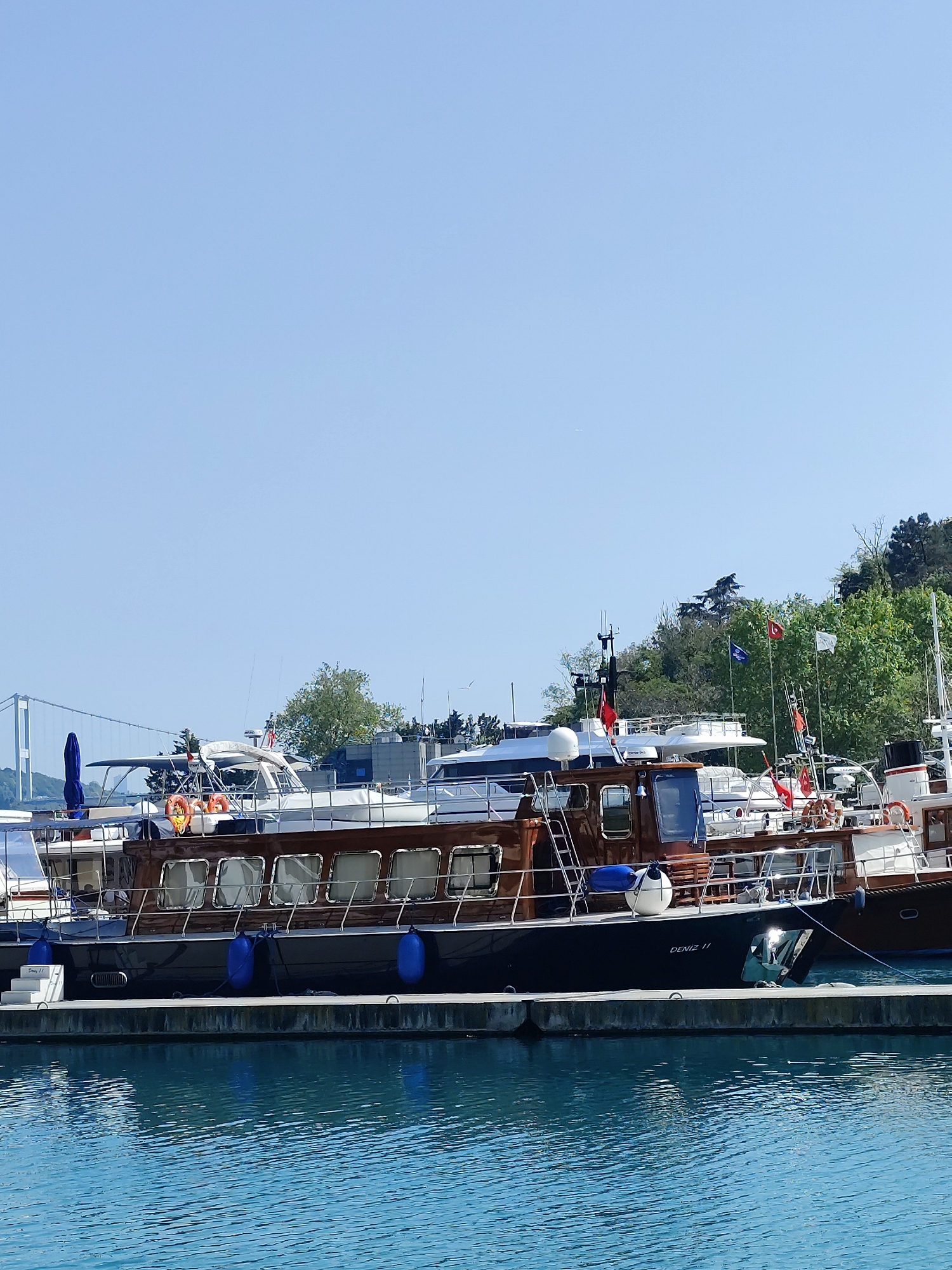 The image shows a marina in Sarıyer, Turkey. Several boats, including one named "Deniz 11", are docked alongside a concrete pier in turquoise water. In the background is the Fatih Sultan Mehmet Bridge and trees. The sky is clear and blue.