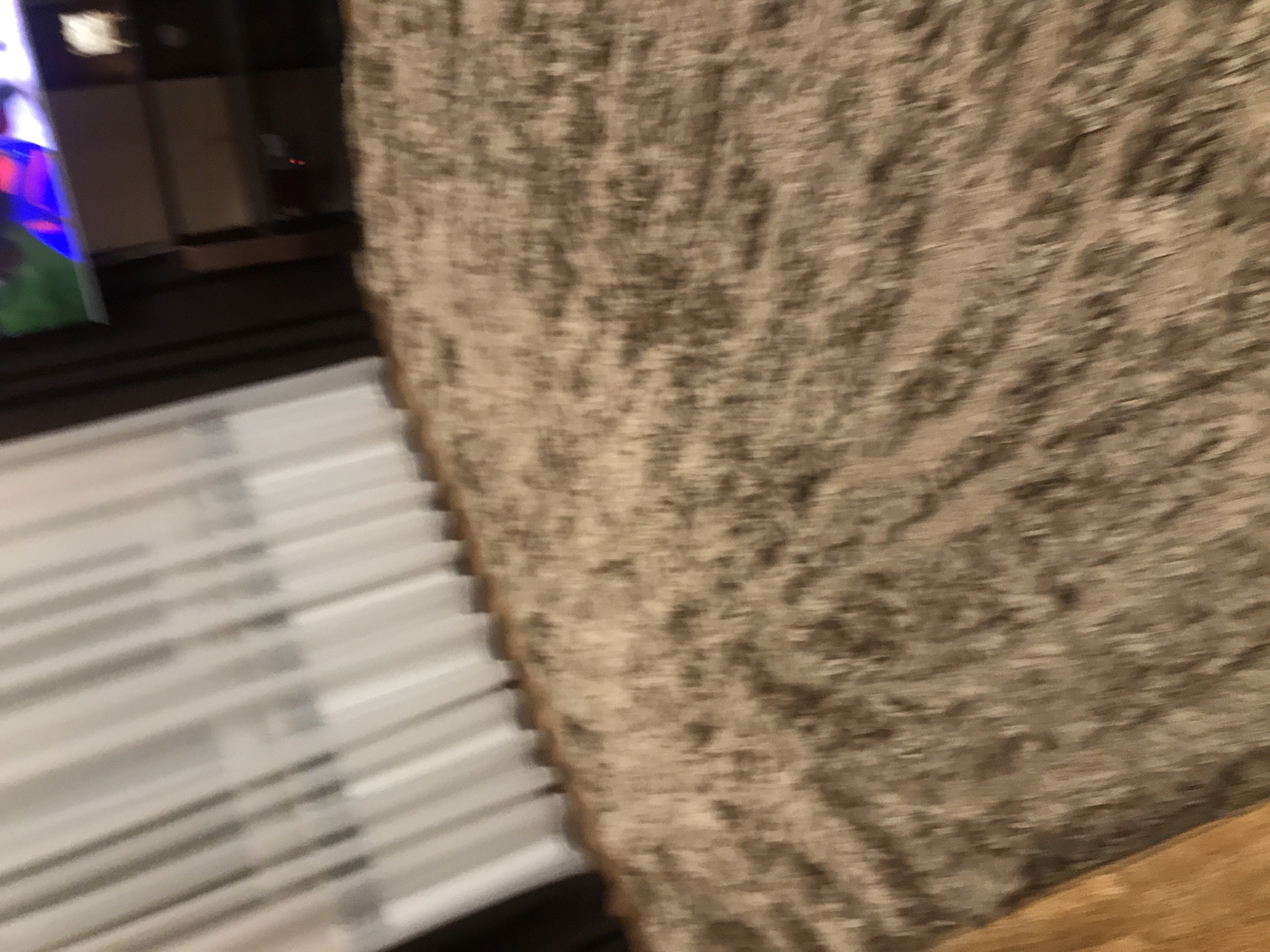 The image shows a blurred view of a room interior. In the left portion, there are white window blinds and what appears to be a dark television or monitor with a colorful image displayed on its screen. To the right, there's a textured wall or surface, possibly carpet or fabric, and a wooden surface at the bottom. The specific location in Yerevan, Armenia is not identifiable from the given visual information.
