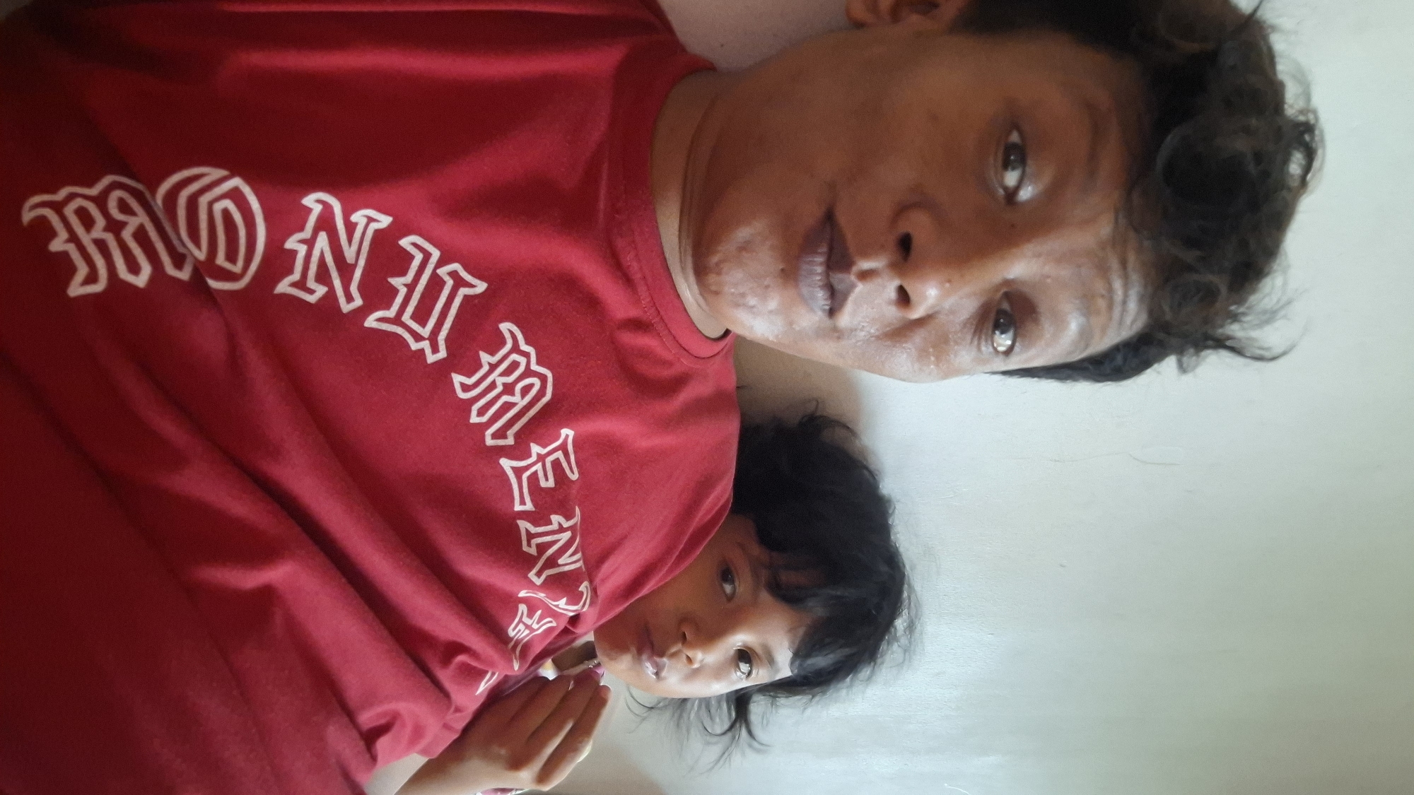 This is a selfie taken indoors, likely in a residential setting in Cibinong Hilir, Indonesia. A person with dark, curly hair is in the foreground, wearing a red t-shirt with white gothic-style lettering. Their face is partially obscured by the camera angle, but their eyes are open and looking forward, and their lips are pursed. Behind them, a child with dark hair is leaning against them, also looking towards the camera. The child's face is partially visible, and they appear to be making a similar facial expression. The background is a plain, light-colored wall. The lighting is soft and diffused.