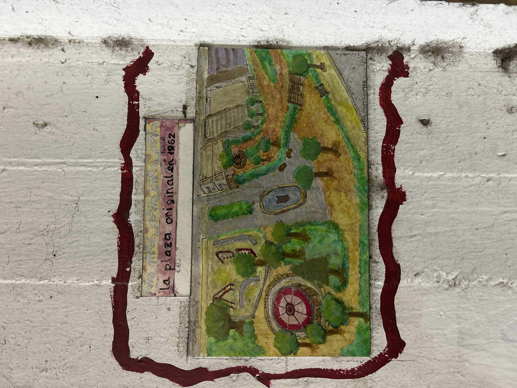 A watercolor painting on a rough, white, concrete wall depicts an aerial view of a park, framed by a dripping, dark red border. The painting is titled "la plaza originalen 1962" in handwritten black script on a decorative strip above the park scene.
The park features various elements:
* A circular structure with a red and white patterned center, possibly a fountain or a small amphitheater.
* A small blue pond with a dark shape inside.
* A playground area with slides and a climbing structure.
* Numerous trees with green canopies.
* Paths and grassy areas.
* Buildings or structures with tiered roofs in the background.
* A wooden structure resembling a bridge or walkway.
The painting is signed at the bottom right with illegible text and the date "1/22 1962". The wall itself shows signs of age and wear, with some scuff marks and texture variations. The painting is attached to the wall, and the red border appears to be painted directly onto the wall. The overall impression is of a historical representation of a public space. This specific artwork is located in the City of Buenos Aires, Argentina, as it is part of a collection of murals in the Saavedra neighborhood that depict historical scenes and places of the area.