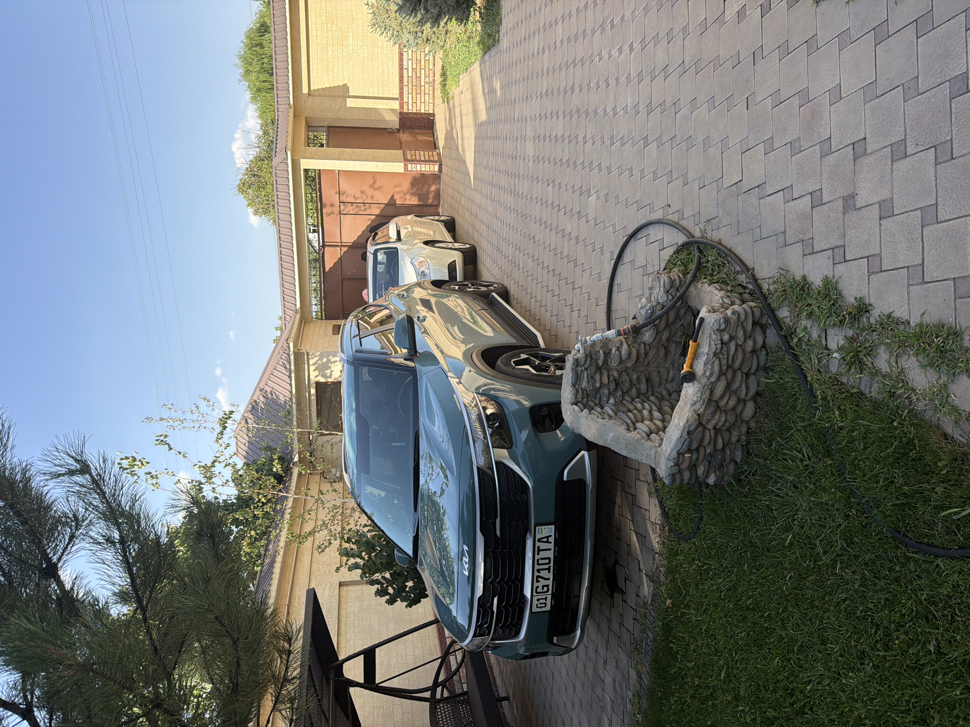 The image depicts a paved driveway in Koshkurgon, Uzbekistan, with two cars parked on it. In the foreground, a teal-colored Kia Seltos is parked, facing forward. To its right and slightly behind, a white car is also parked, facing the same direction. The driveway is constructed of interlocking beige pavers. To the left of the Kia, a black metal swing set with a bench is visible. Beyond the cars, a large brown metal gate stands, flanked by a light yellow brick wall. The scene is set against a backdrop of green trees and a clear blue sky with a few scattered clouds. A decorative stone structure, resembling a small well or fountain, is present in the foreground to the left, with a black hose coiled around it. The sun is casting shadows, indicating it is likely daytime.