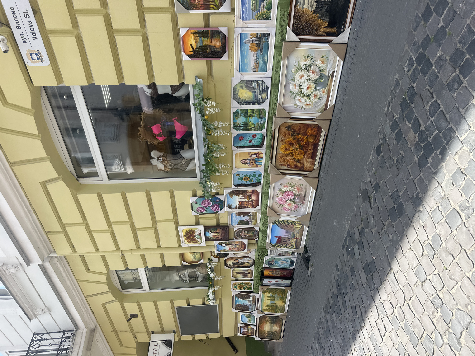 The image depicts an outdoor art exhibition in Ternopil, Ukraine, on Valova Street. A variety of framed paintings are displayed on the cobblestone sidewalk in front of a pale yellow building with a large window. The building facade features decorative brickwork and architectural moldings.
The paintings showcase a range of subjects, including landscapes, still lifes, and figurative art. Prominently featured are floral arrangements, depictions of natural scenery with water and trees, and scenes with architectural elements. Some smaller paintings are stacked vertically on top of larger ones.
Through the building's window, mannequins are visible, dressed in lingerie, including a pink bikini set. A sign above the window on the building reads "вул. Валова" and "Valova St.", identifying the location. The street is paved with dark grey cobblestones, with a shadow cast across the lower portion of the image. The overall scene suggests a commercial street with a focus on art sales. No people are directly visible in the image.