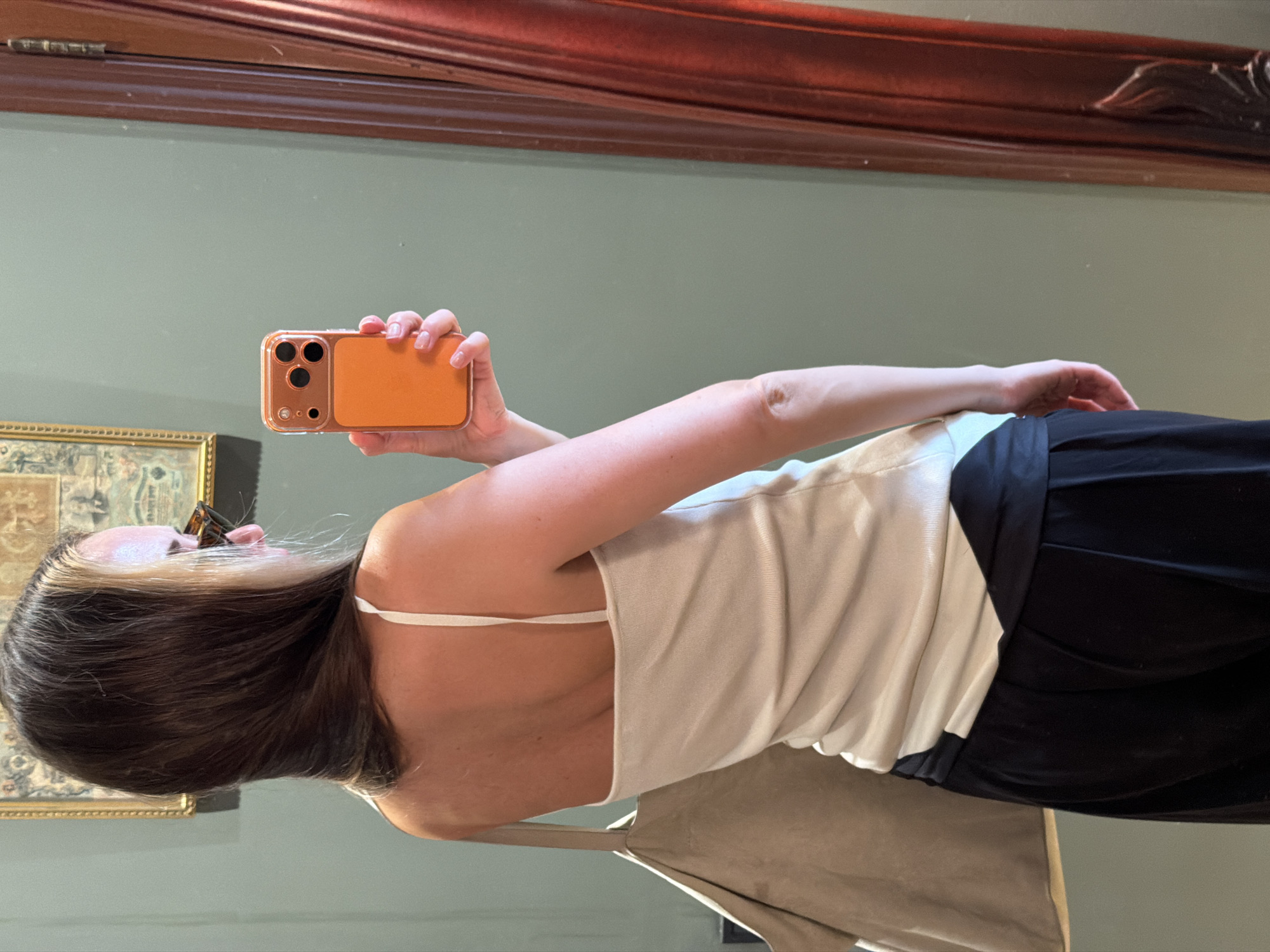 A woman with short, dark hair and a blonde money piece is taking a selfie in a mirror. She is wearing a white spaghetti-strap tank top and black pants. She is holding an orange iPhone in her right hand. The mirror is reflecting a room with a green wall and a dark wood trim along the top. There is a framed painting on the wall to the left. The location is not possible to determine from this image.