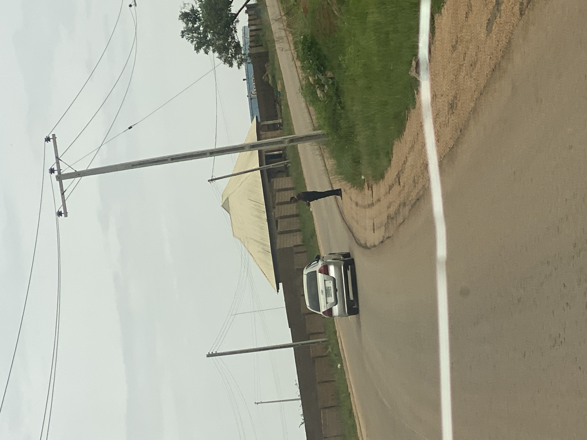 A silver hatchback car is driving on a paved road in a rural area. The car is facing away from the viewer and is in the center of the road. A person is standing on the side of the road next to the car. They are wearing dark clothing and appear to be facing the car.
The scene is an outdoor setting with a cloudy sky overhead, suggesting it could be morning or late afternoon. The road is lined with grass and some vegetation. There are utility poles with wires running overhead, indicating the presence of electricity infrastructure. In the background, there are buildings with light-colored roofs and brick walls. One building appears to be a large structure covered by a light-colored awning or tarp. The overall impression is a somewhat overcast day in a developing area, possibly a town or village. There is a white line painted on the road, marking the lane. No other specific location cues are immediately apparent beyond the general rural setting in DU, Nigeria. The visible text on the car's license plate is obscured. The person standing by the car has their back to the camera, making it impossible to discern any emotional expression.