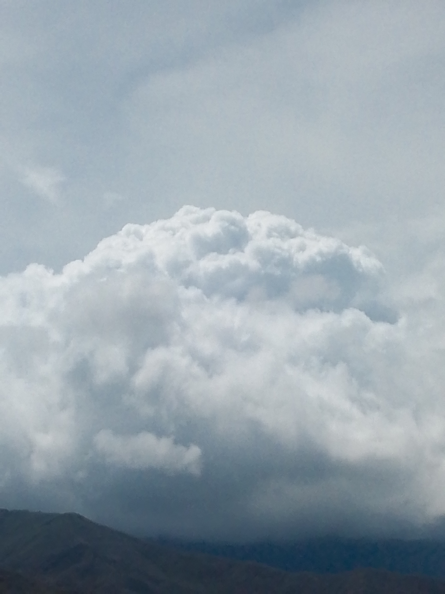 The image shows a large, puffy cumulonimbus cloud formation in a light blue sky. The cloud dominates the view, appearing dense and white with varying shades of gray where the sunlight is obscured. At the bottom of the frame, a dark, indistinct mountain range or elevated terrain is visible, likely at some distance due to the hazy atmosphere. No people or other objects are evident. The location is not determinable as Juan Griego, Venezuela, based on the image alone.
