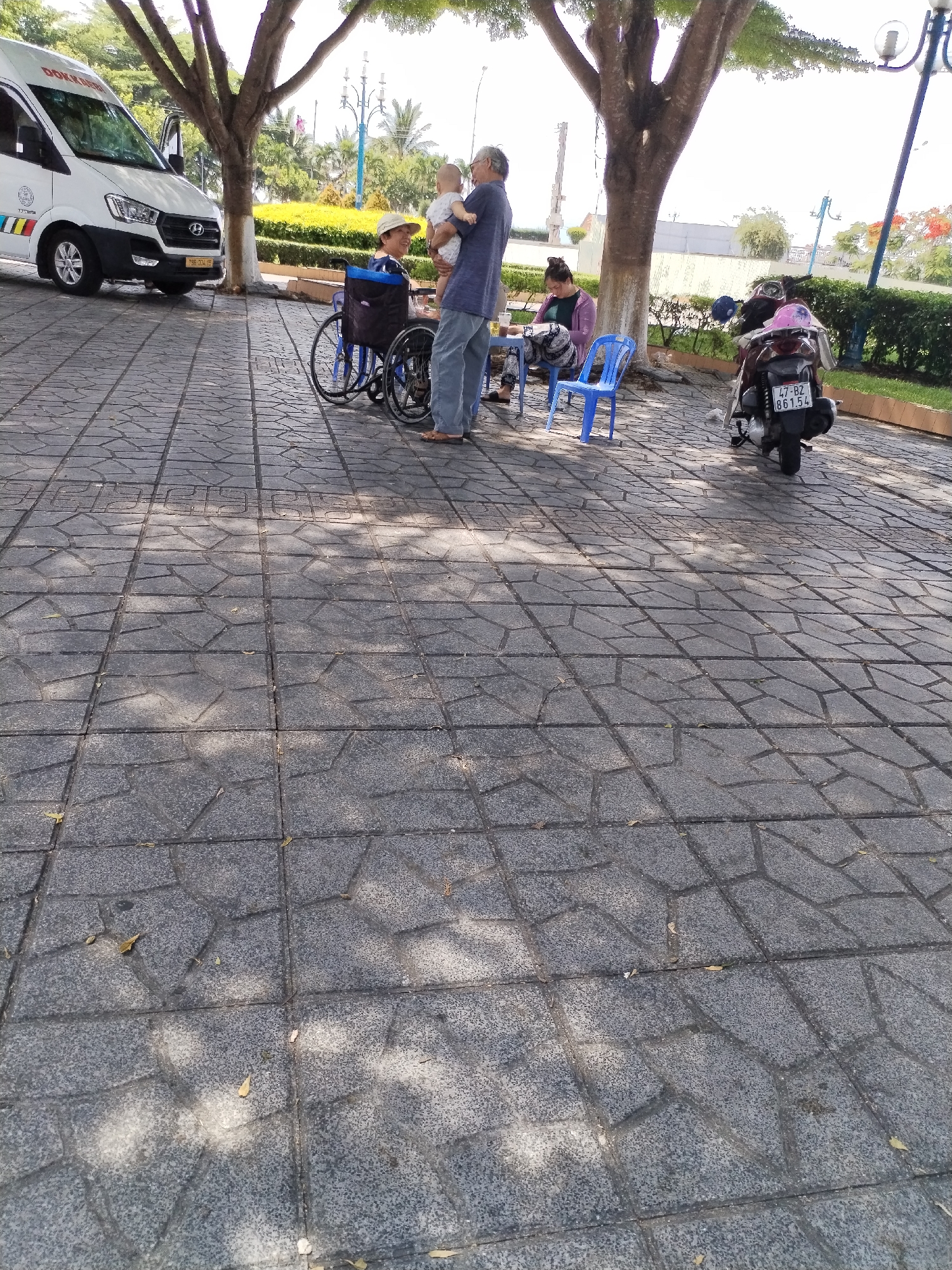 In the city of Bac Nha Trang Ward, Vietnam, on a paved stone walkway shaded by trees, a scene unfolds involving several individuals and vehicles. A white van, possibly an ambulance due to its markings, is parked on the left side of the frame. Nearby, a man in a blue shirt and jeans holds a baby, while a woman in a wheelchair wearing a hat smiles at them. Further to the right, another woman is seated at a small table under a tree, and a blue plastic chair is placed in front of her. A parked motorcycle, with a license plate reading "47-BZ 861.54," is visible in the background, with a pink helmet on its seat. The environment suggests a public outdoor space, possibly a park or a communal area.