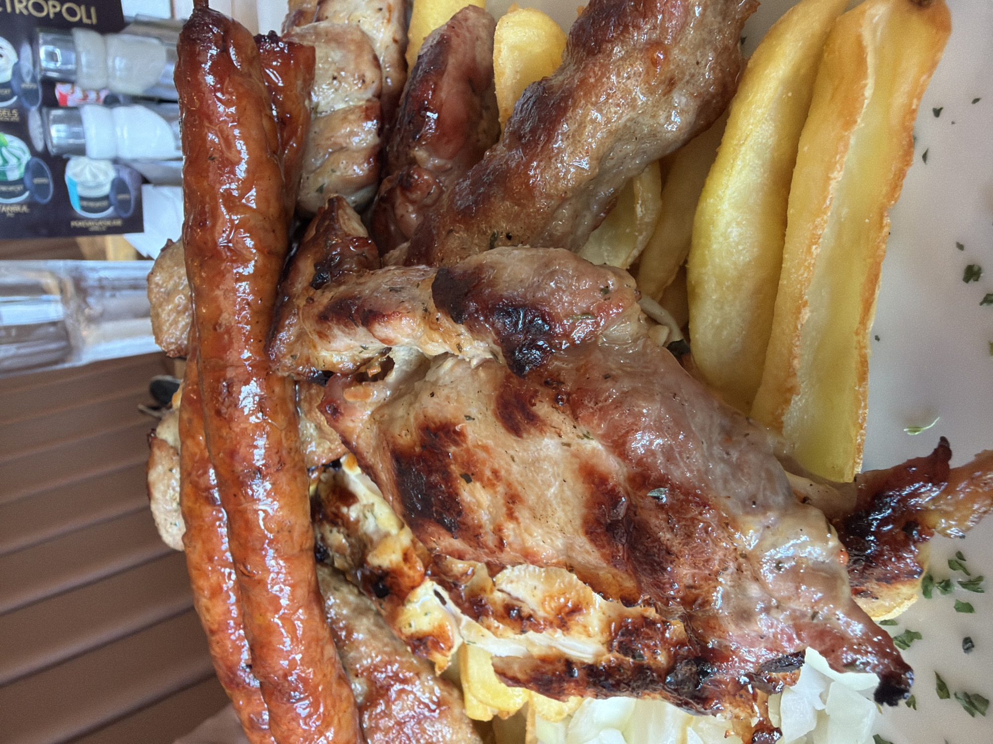 A close-up, overhead shot of a platter of grilled meats and fries is presented. The platter contains several pieces of grilled meat, including what appear to be sausages and chicken or pork cutlets, arranged on a bed of thick-cut French fries. The meats show signs of charring and glistening from the cooking process, with visible herbs sprinkled on some pieces. Underlying the meats, a scattering of finely chopped white onion is visible. In the background, a clear glass of water and what appears to be a menu or advertisement with some bottles are out of focus. The lighting is bright, highlighting the textures and colors of the food. The setting appears to be an outdoor or casual indoor dining area, with a wooden-textured surface visible at the top of the frame. The image was taken in the vicinity of Bigor, Montenegro.