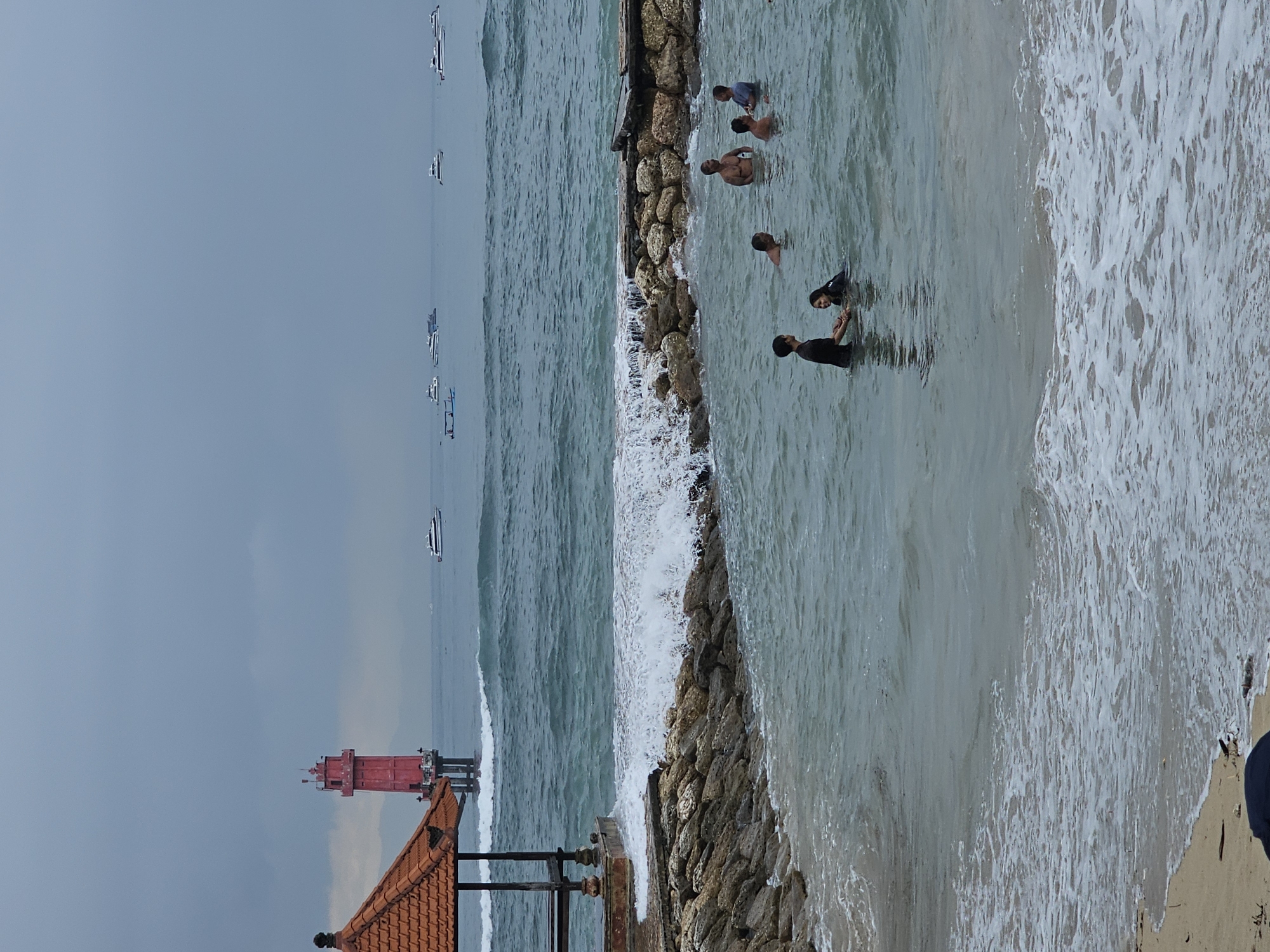 The image captures a seascape with a rock breakwater partially dividing the scene. The sky is overcast, suggesting cloudy weather. In the distance, there are boats on the water.
In the foreground, a structure with a red tile roof and a red tower-like structure, possibly a lighthouse, is partially visible.
To the right of the breakwater, several people are in the water, some standing and some submerged to their shoulders. The water is churning with waves hitting the breakwater and washing onto the shore.
The scene is from the city of Denpasar, Indonesia. The exact location within Denpasar is not discernable from this image.
