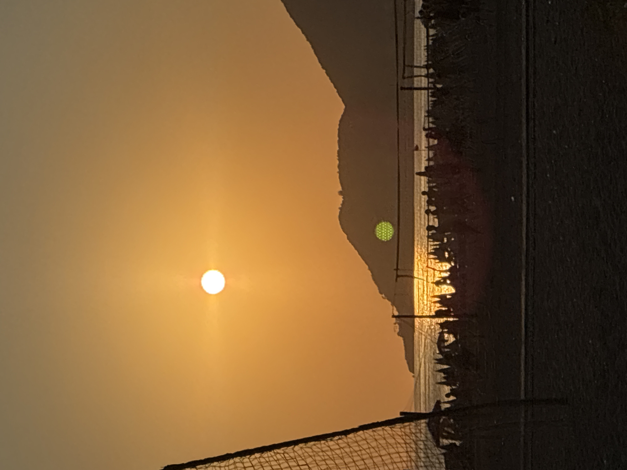 The image depicts a sunset over the sea, viewed from a beach in Alanya, Turkey. The sun, a bright white orb, is positioned in the upper center of the frame, casting a warm orange glow across the sky. In the foreground, a black volleyball net is partially visible on the left side. The middle ground shows a silhouette of a beach with numerous people and umbrellas, and a body of water reflecting the sunset. Hills form the background, silhouetted against the vibrant sky. There is a green hexagonal glare on the left side of the hill.