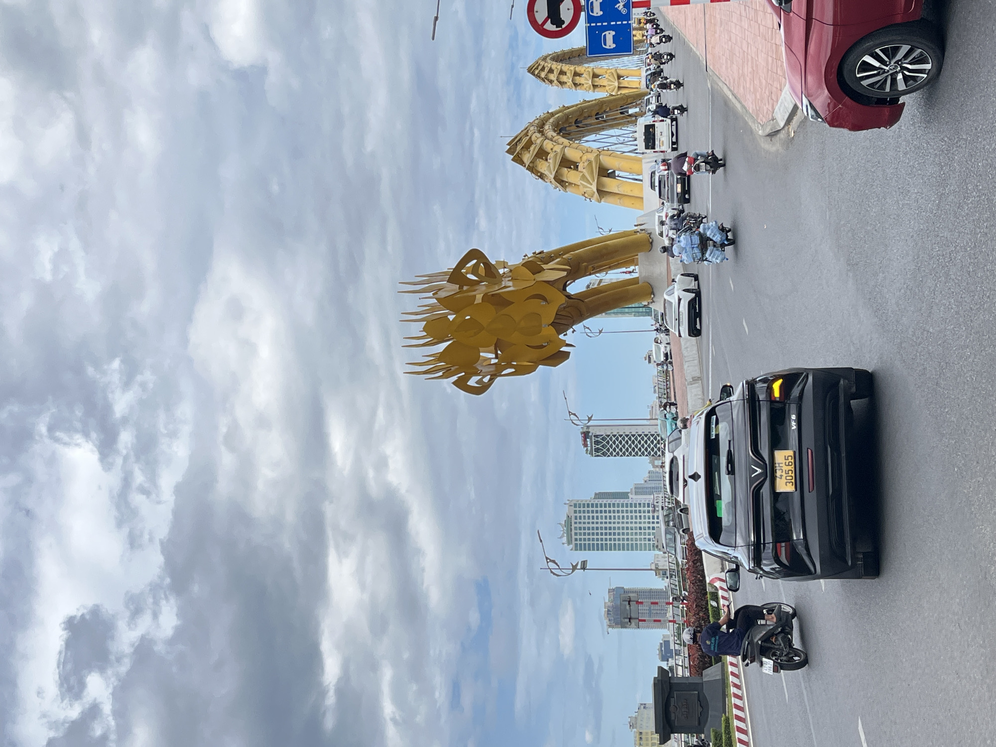 The image displays a bustling urban street scene in Đà Nẵng, Vietnam, featuring the iconic golden Dragon Bridge under a cloudy sky. The photograph, originally oriented vertically, shows a multi-lane road running horizontally in the foreground and midground, with the bridge spanning across the midground.
The most prominent subject is the Golden Dragon Bridge, with its elaborate, spiky-maned dragon head visible on the left and its arched, golden body extending towards the right. Above the bridge, several road signs are mounted. One blue rectangular sign features white pictograms of a car, motorcycle, bus, and bicycle, indicating allowed vehicle types. Adjacent to it, a red circular sign with a diagonal bar over a truck pictogram clearly prohibits trucks.
The road is active with various vehicles. In the foreground, a black VinFast VF 6 SUV is visible from the rear, displaying the license plate "43H 305.65". A person wearing a helmet and a blue shirt rides a black motorcycle directly behind the SUV. On the far right, the front bumper and wheel of a red car are partially visible. Further down the road, more motorcycles and cars are navigating traffic, including a motorcyclist remarkably carrying multiple large water bottles. A white truck or van is also present in the vehicle line-up.
In the background, beyond the bridge and traffic, a skyline of modern high-rise buildings rises under a sky filled with grey and white clouds, indicating a daytime setting. The overall scene captures the vibrant energy of Da Nang's urban environment.