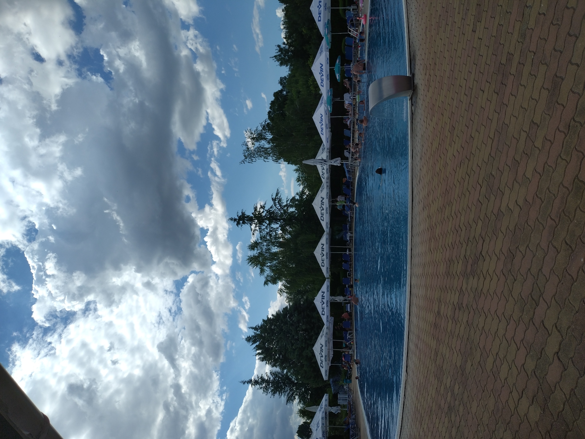 The image shows an outdoor swimming pool in Yagoda, Bulgaria. The pool is filled with blue water and surrounded by a paved area with brown interlocking bricks. Several white umbrellas with the "Devin" logo are lined up around the pool, providing shade. Beneath the umbrellas are blue lounge chairs, some of which are occupied by people. The pool itself has several people swimming and relaxing in it. A modern, silver waterfall feature is visible on the edge of the pool. In the background, there are trees and a cloudy sky. The overall scene depicts a recreational area on a partly cloudy day.