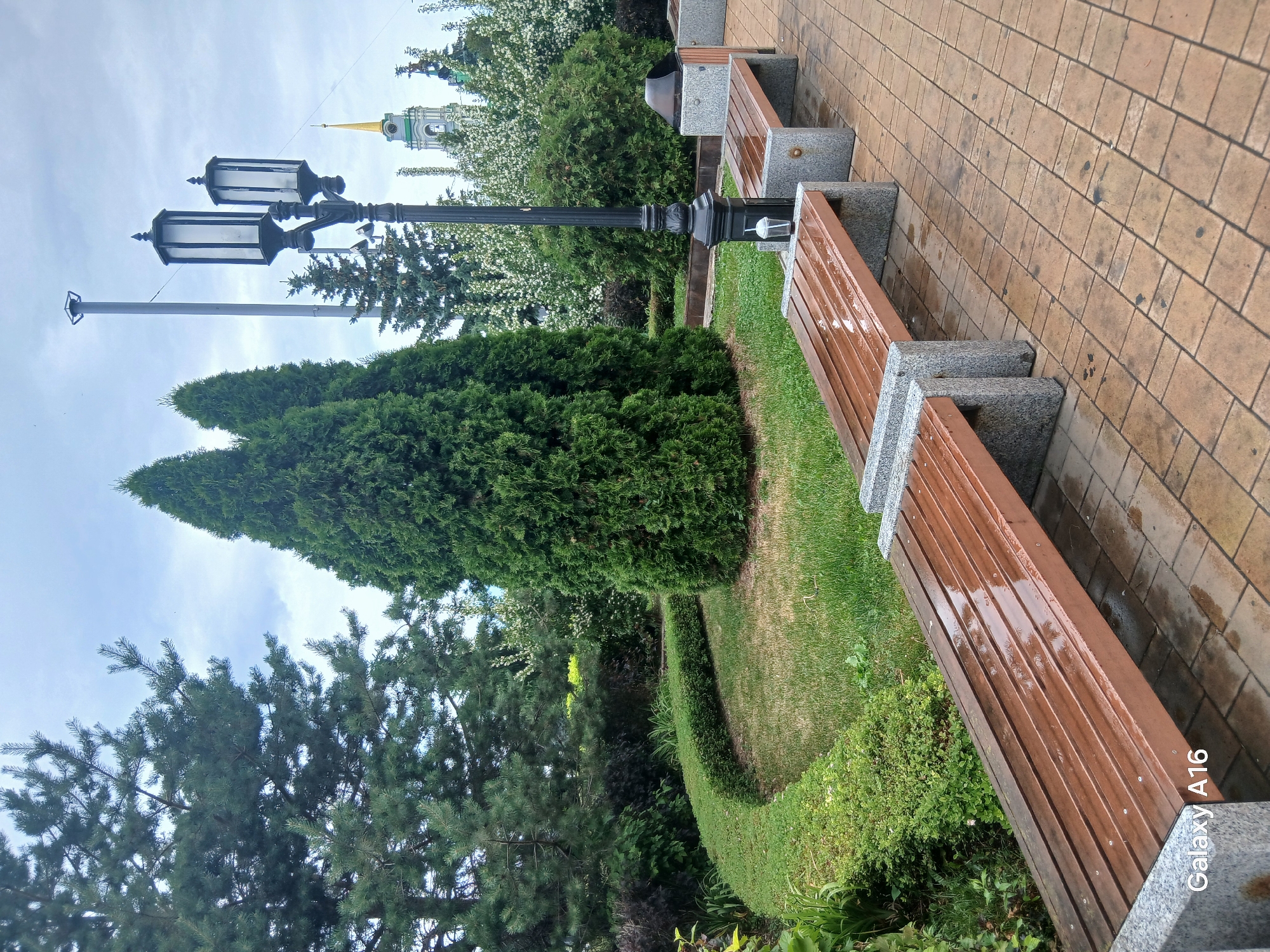 The image displays a park scene with a paved walkway and two wooden benches. The walkway is made of brown rectangular bricks laid in a pattern, with visible dark spots, possibly from moisture. The benches are made of wooden slats and supported by granite-like bases. The park is green with manicured hedges and various trees, including evergreens and flowering bushes. A lamppost with two lights stands near the benches. In the background, a church or tower with a green and gold spire is visible among the trees, suggesting a historical or significant location. The sky is overcast, and the presence of water on the benches and walkway indicates recent rain. The overall mood is serene and perhaps slightly damp due to the weather. The image also contains the text "Galaxy A16" in white lettering on the bottom right.