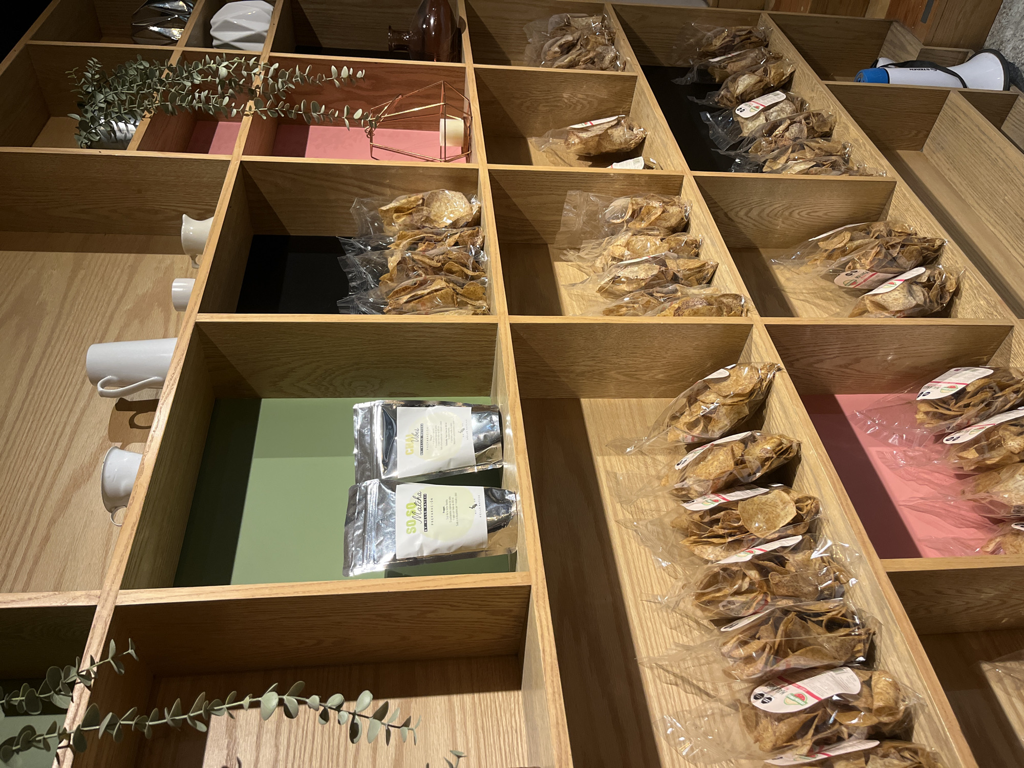 A wooden shelving unit displays various items. The unit contains decorative plants, ceramic mugs, bags of chips, and tea packets. One shelf holds a white megaphone. The interior of some shelves is painted in pink, green, and black. The location is likely a shop or store, but the city of San Bernardino Tlaxcalancingo, Mexico cannot be definitively determined from the image. There are no visible people or interactions in the image.