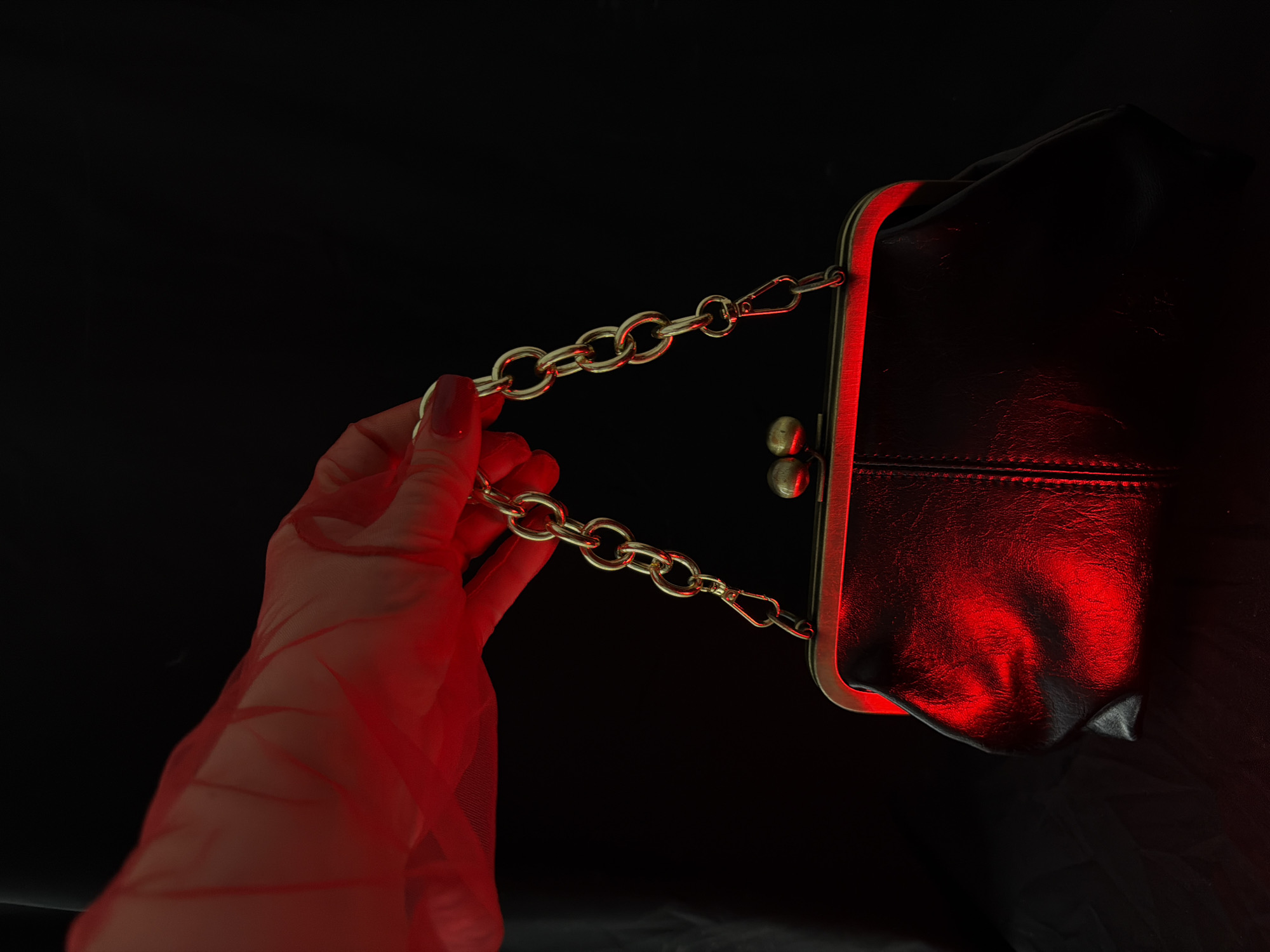 A close-up, low-angle studio shot shows a hand wearing a sheer red glove holding a dark-colored, framed purse with a gold chain strap. The purse is lit with a dramatic red light that casts a glow on its textured surface. The background is black.