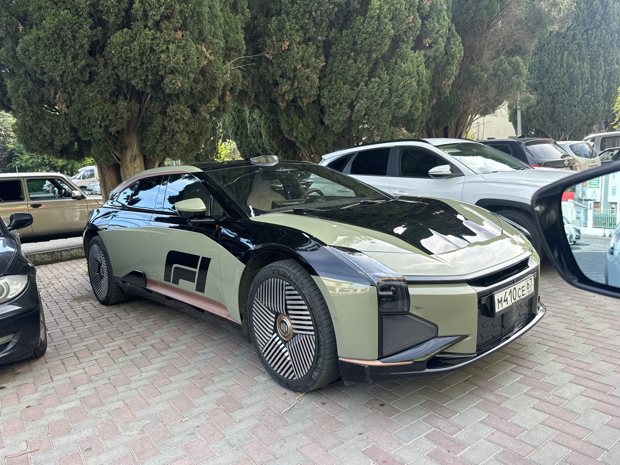 The image features a distinctive two-tone HiPhi Z electric vehicle prominently parked on a paved surface, likely in Sochi, Russia. The car, the primary subject, boasts a futuristic design with a black upper section and a light green or khaki lower body, accented by subtle rose gold trim along the side skirts and window frames. Its large, multi-spoke wheels exhibit an intricate, aerodynamic pattern, with a central "HiPhi" logo. A black dome-shaped sensor is visible on the roof. On the lower front door, a stylized "R" or "FI" graphic is present.
The car's front license plate, "M410 CE 67 RUS," is clearly visible, with "AVILON" inscribed below the main registration number, confirming its Russian context. The vehicle is parked outdoors on light-colored, reddish-brown cobblestones or pavers.
In the background, dense, dark green coniferous trees, resembling cypresses, form a lush, natural backdrop, suggesting a tree-lined street or park setting. Other vehicles are partially visible around the main car: part of a black car to its immediate left, a beige sedan further left, and a white SUV (possibly a Hyundai) behind it, along with other indistinct vehicles in the distance.
The scene is set during daytime under bright, clear weather conditions. No people are visible, and no specific activity is occurring beyond the cars being parked. The overall impression is of a modern electric vehicle situated in a verdant, urban environment.