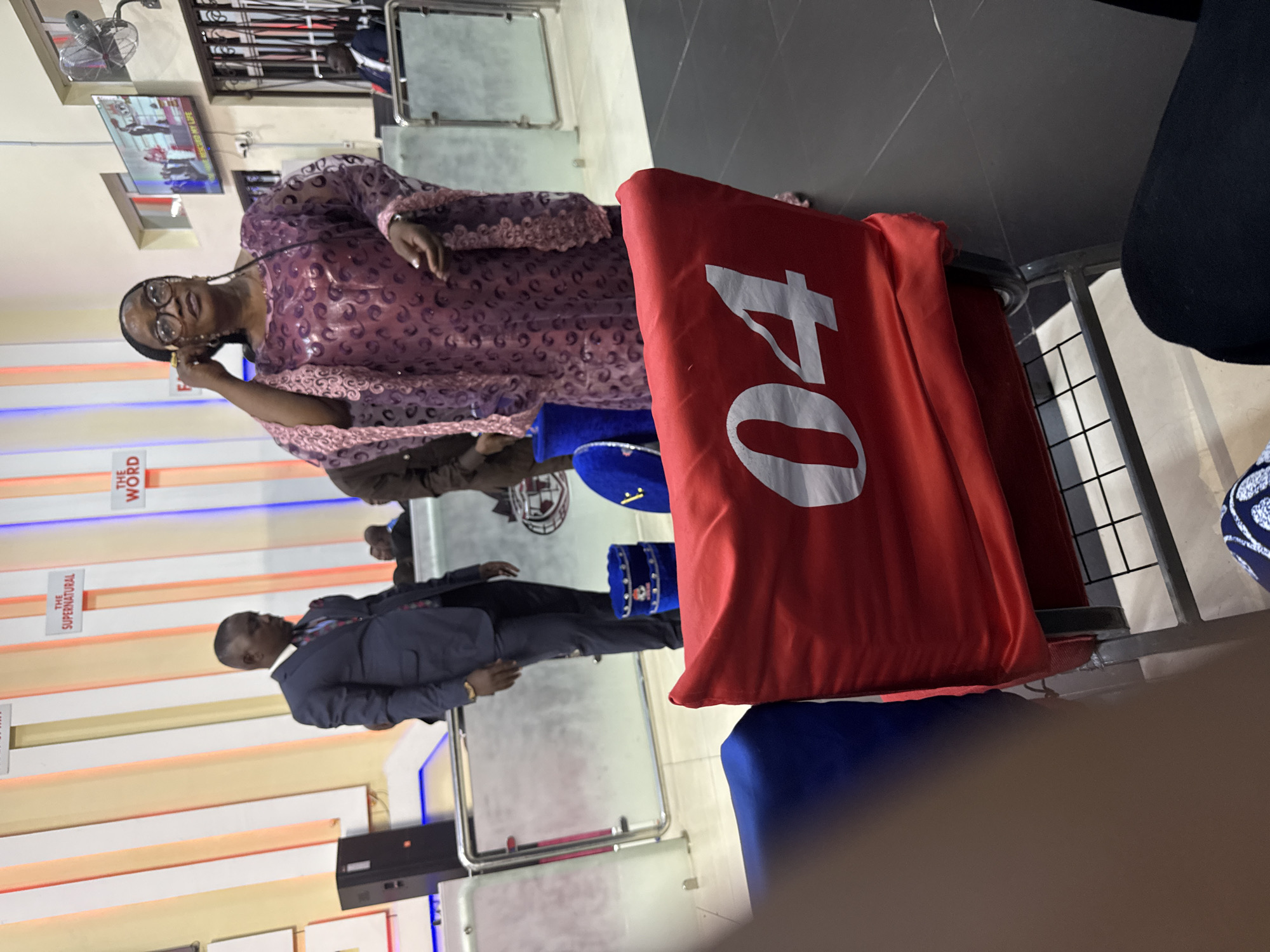 The image shows a room with multiple individuals. A woman wearing glasses and a patterned dress is holding something near her ear, possibly a phone. A man in a suit stands near a wall adorned with vertical signs. There's a red covering with the number "04" prominently displayed, and other people are seated in the background. The location is Iddo Tudunwada, Nigeria.