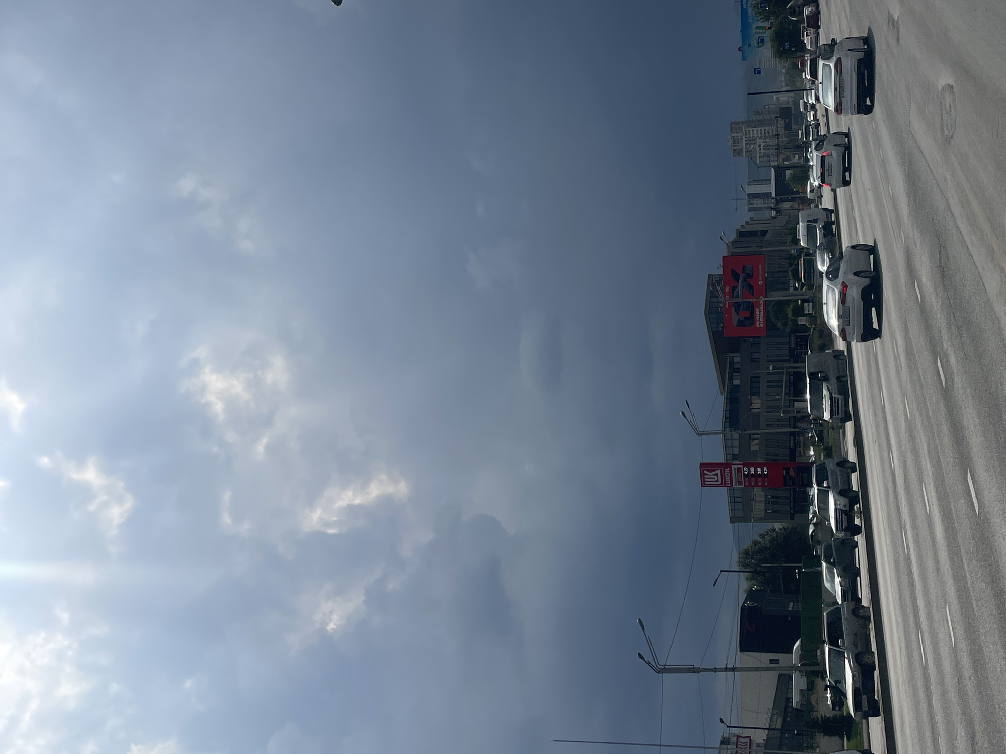 A street scene in Tbilisi, Georgia, on a cloudy day. The sky is filled with dark, heavy clouds with rays of sunlight breaking through in the upper left quadrant. A multi-lane road with light traffic dominates the lower half of the image. Several cars are visible on the road, moving in both directions. On the right side of the road, there are buildings, including a Lukoil gas station with a prominent red sign displaying fuel prices. Streetlights and utility poles line the side of the road. In the background, more buildings and a distant cityscape are visible. The overall impression is one of an urban environment under an impending or passing storm.