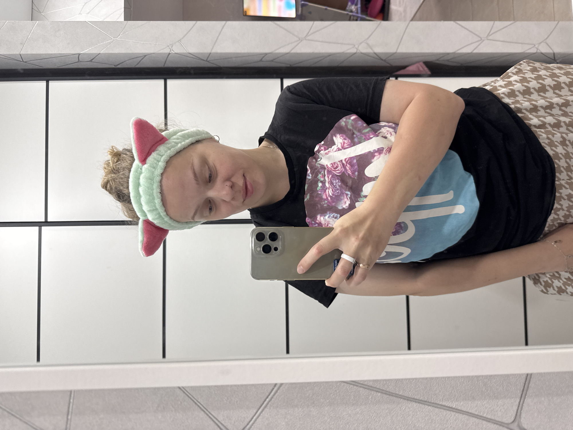 In Mikhaylovsk, Russia, a young woman is taking a selfie in a mirror. She is wearing a black t-shirt with a floral design and a light brown and white patterned skirt. She has a mint green headband with pink cat ears on her head. In her right hand, she is holding a smartphone, and she is wearing a white ring and a gold ring on her left hand. The background shows a white closet with black horizontal lines, and a textured wall with a geometric pattern. A television screen is visible in the reflection on the right side of the image.