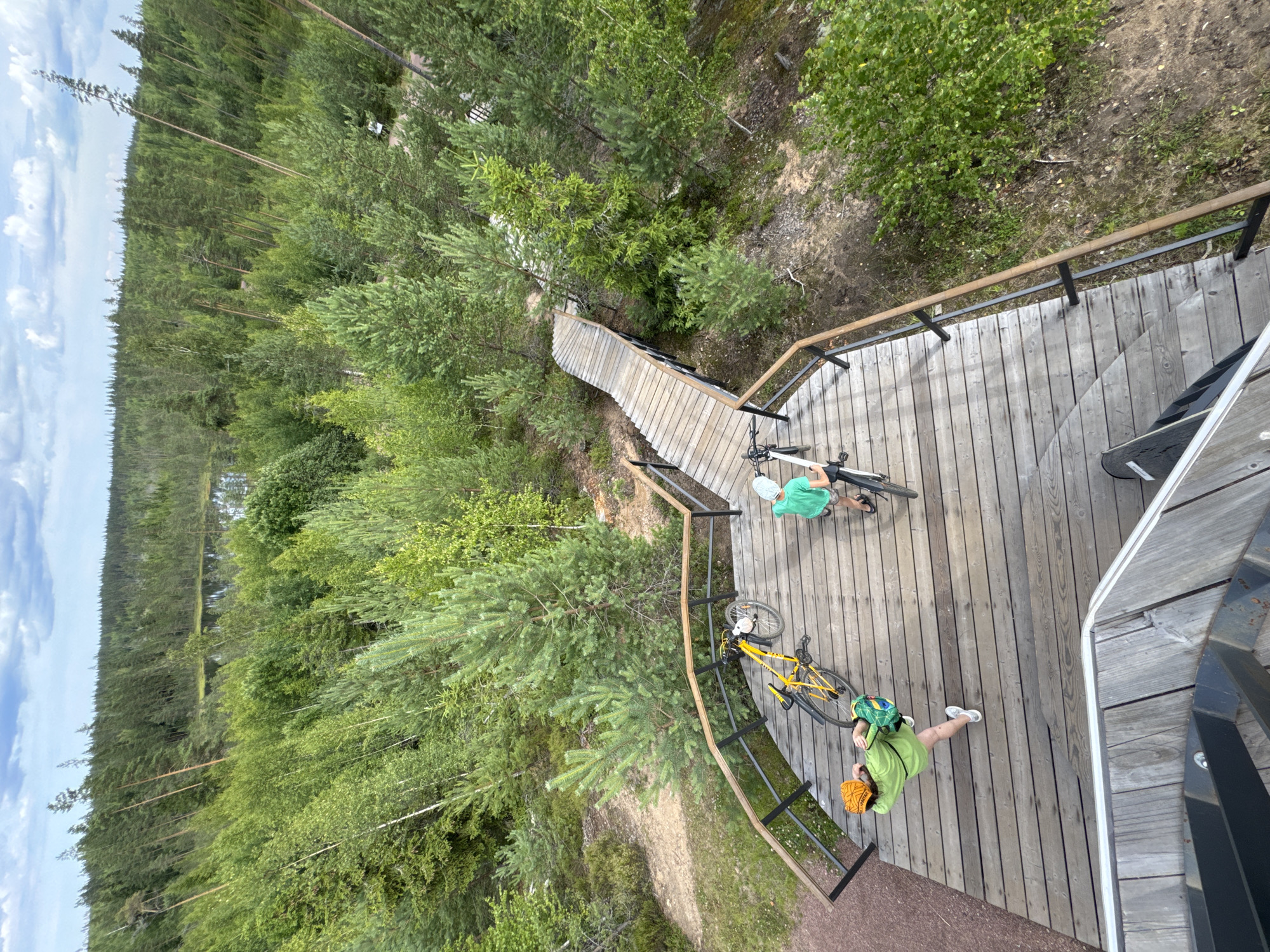 An overhead perspective shows a wooden viewing platform with two individuals and bicycles. On the left, a person wearing a green shirt and an orange helmet is leaning over a yellow bicycle. To the right, a person in a green t-shirt and a light-colored hat stands next to a bicycle. Both individuals are on the wooden platform, which features a railing with dark posts and wooden handrails. A winding wooden staircase extends from the platform into a dense forest area. The surrounding environment is a forest with various trees, including pines and birches, and a body of water is visible in the distance. The sky above is cloudy. The location is the city of Peski, Russia.