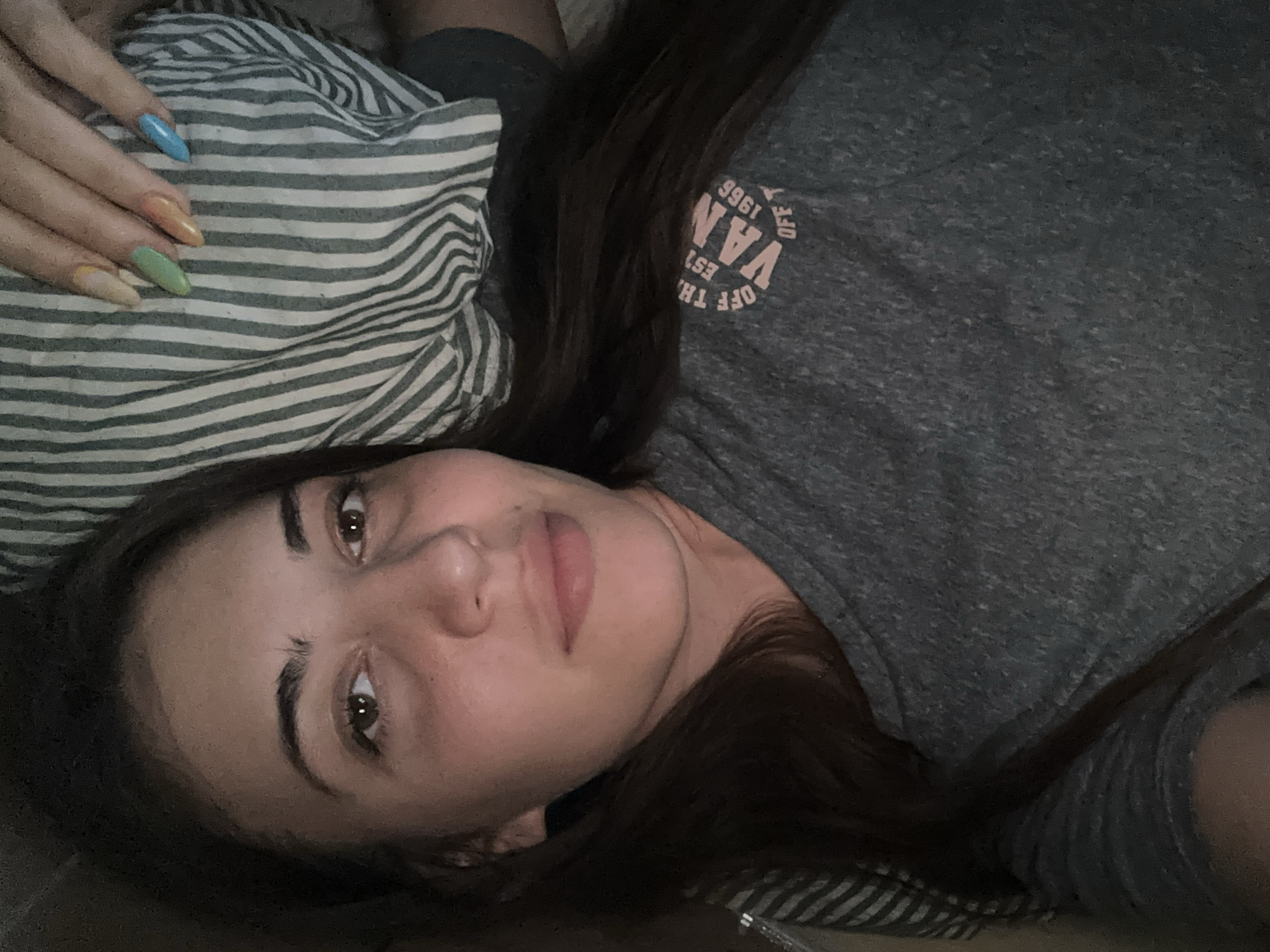 A young woman is depicted lying down indoors, looking directly at the camera with a relaxed, neutral expression. Her dark brown hair is spread out around her head. She has brown eyes and dark eyebrows. She is wearing a grey t-shirt that features a circular pink logo on the chest. The logo reads "OFF THE WALL" at the top, "VANS" in the center, and "EST. 1966" below, all enclosed within the circle.
Her left hand, adorned with long, almond-shaped nails painted in a variety of pastel colors including blue, orange, green, and a light yellowish-beige, rests on a striped pillow or blanket beside her head. The bedding displays a pattern of thin grey and white stripes. The lighting is soft and ambient, indicating an indoor setting, likely in the evening or low-light conditions. The overall mood of the image is calm and reposeful. There are no specific visual cues within the image itself to identify the location as Sirakovo, Bulgaria.