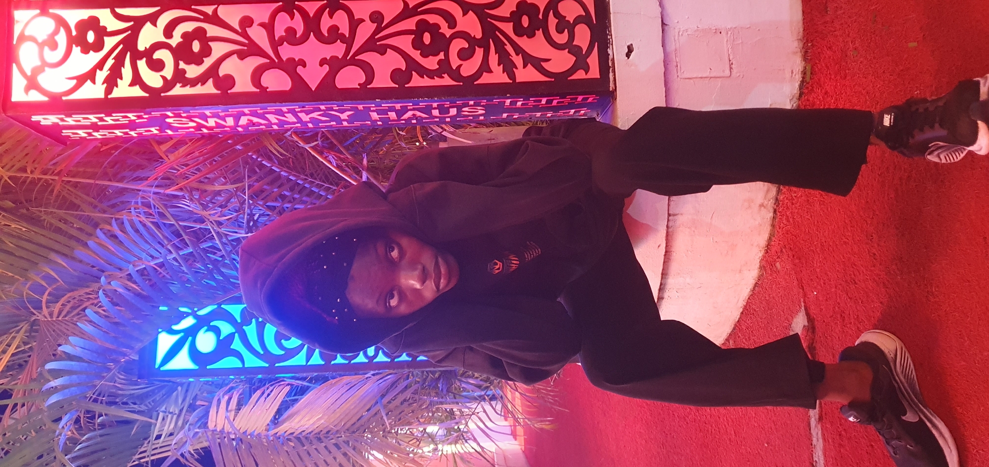 A young woman with dark skin is seated on a low, white, circular structure in front of a decorative, illuminated column and palm fronds. She is wearing a black hooded sweatshirt and black pants, with her hands on her head inside the hood. The column behind her is lit with pink and red hues on the right and blue on the left, with an ornate, cut-out design. The ground is covered in a bright red carpet-like material. The woman is looking directly at the camera with a neutral expression. The scene appears to be an outdoor setting at night, possibly a garden or public space in Bar Jirgi Summa, Nigeria, illuminated by decorative lighting.