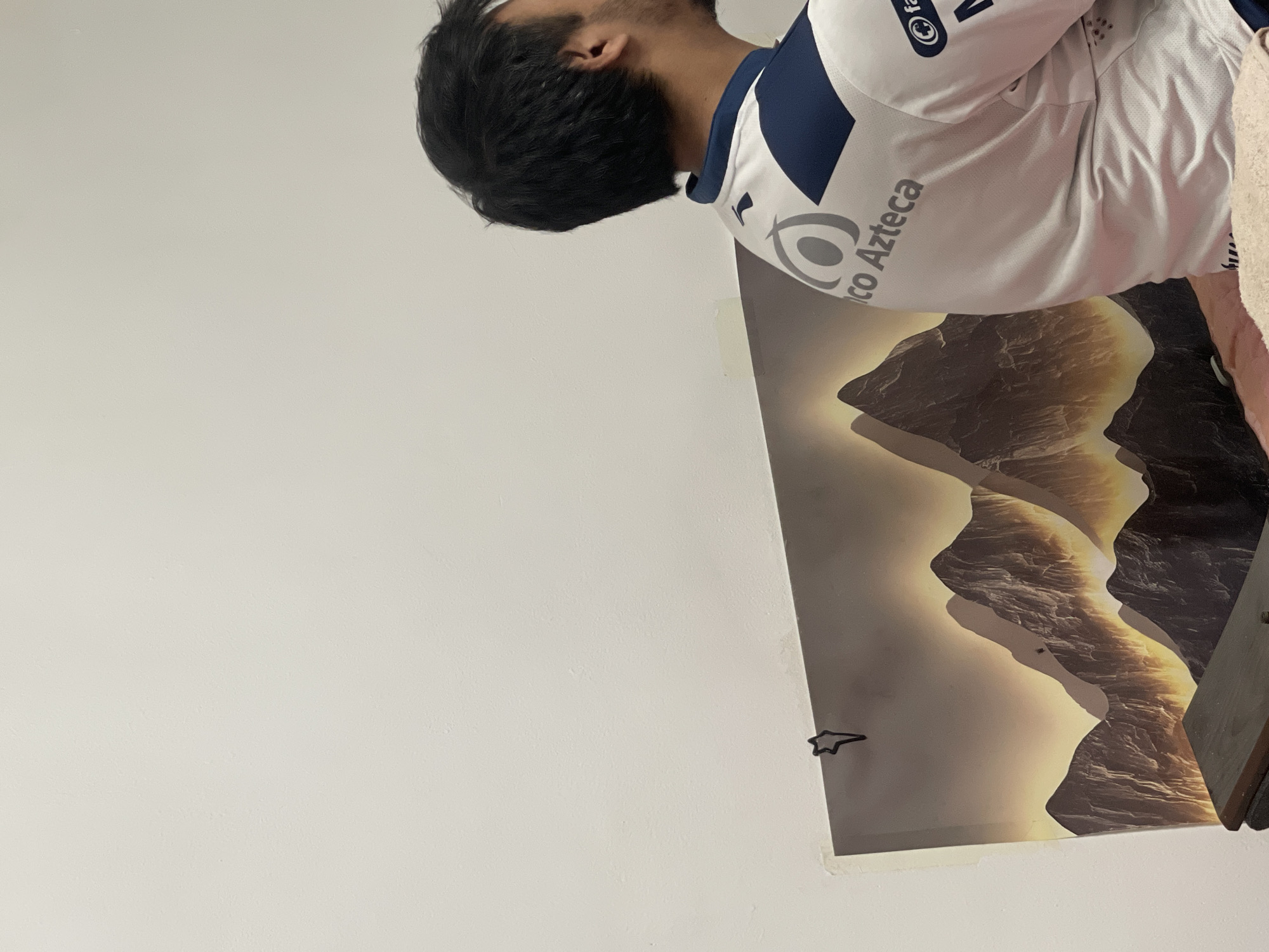 The image depicts the back of a person wearing a white and navy blue polo shirt with the logo "Banco Azteca" visible on the back. The person has dark hair and is facing away from the camera, towards the right side of the frame. In the background, a poster or artwork of a mountainous landscape with a warm, sunset-like glow is displayed on a white wall. A black star-shaped paperclip is attached to the top left corner of the artwork. A dark-colored piece of furniture, possibly a table or cabinet, is visible in the bottom left corner of the image, partially obscuring the artwork. The overall environment appears to be indoors, with plain white walls. The scene does not provide any specific indicators of its location within Bishkek City, Kyrgyzstan.