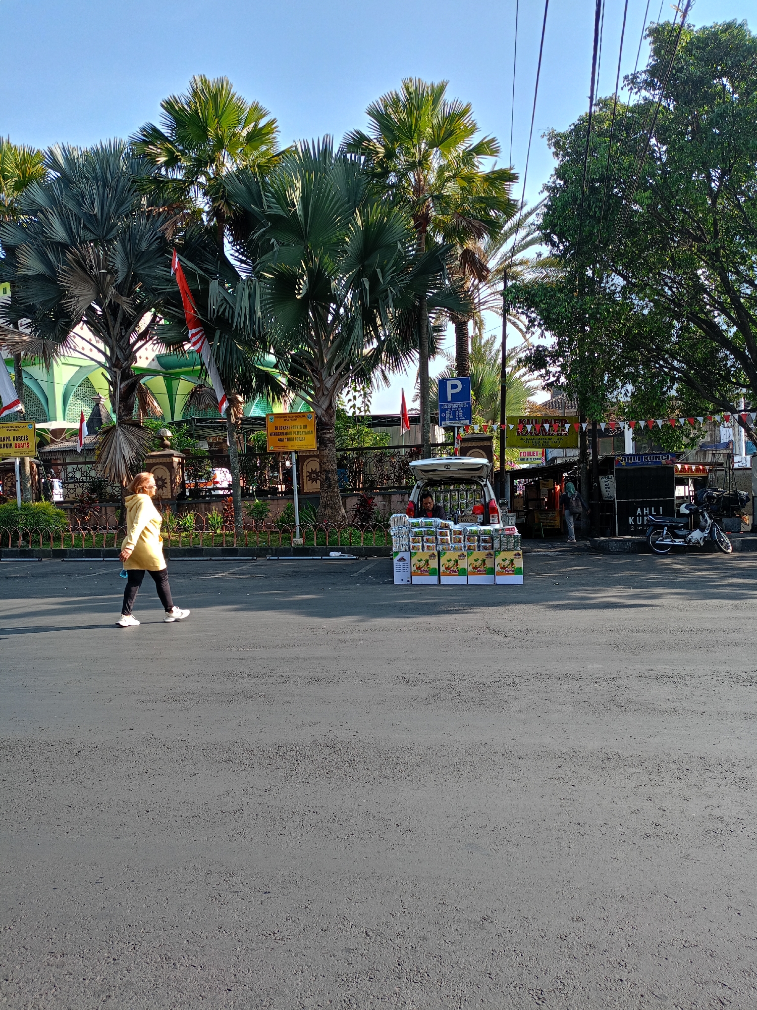 The image depicts a street scene in the city of Batu, Indonesia, on a clear, sunny day. In the foreground, a woman wearing a yellow jacket and black leggings walks across an asphalt road. She is carrying a blue object in her left hand. Behind her, a vehicle with its trunk open displays stacks of boxed goods, possibly food items or products for sale. To the right of the vehicle, a small food stall with a sign reading "KULINER KAUMAN" and "TOILET" is visible. Further to the right, a motorcycle is parked. In the background, palm trees and a building with green accents, likely a mosque, are prominent. Several flags and banners are displayed along the street. The overall atmosphere suggests a bustling urban environment, possibly a market area.