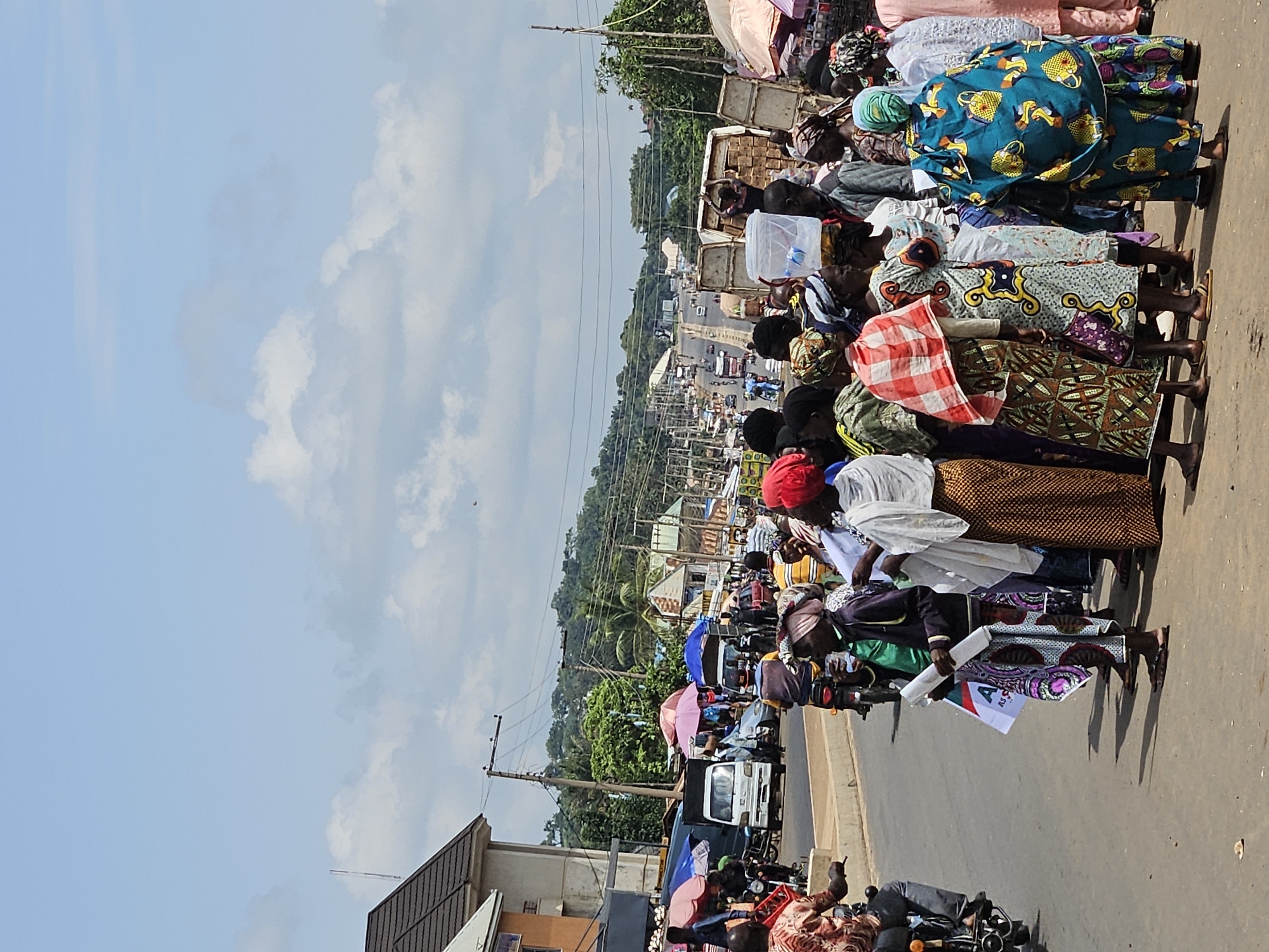 The image depicts a street scene in Akwanga, Nigeria, with a group of people gathered on the side of a road. The people are dressed in colorful clothing, and some are holding items. Several vehicles, including cars, motorcycles, and trucks, are visible on the road, suggesting a busy urban environment. Buildings and market stalls line the street, indicating a commercial area. The sky is partly cloudy, and the overall scene appears to be during the daytime. The people are standing and interacting, with some appearing to be engaged in conversation or observing something. The presence of market stalls and vehicles suggests a lively local economy and transportation hub. The ground is paved, and some debris is visible. The image captures a moment of everyday life in Akwanga.
