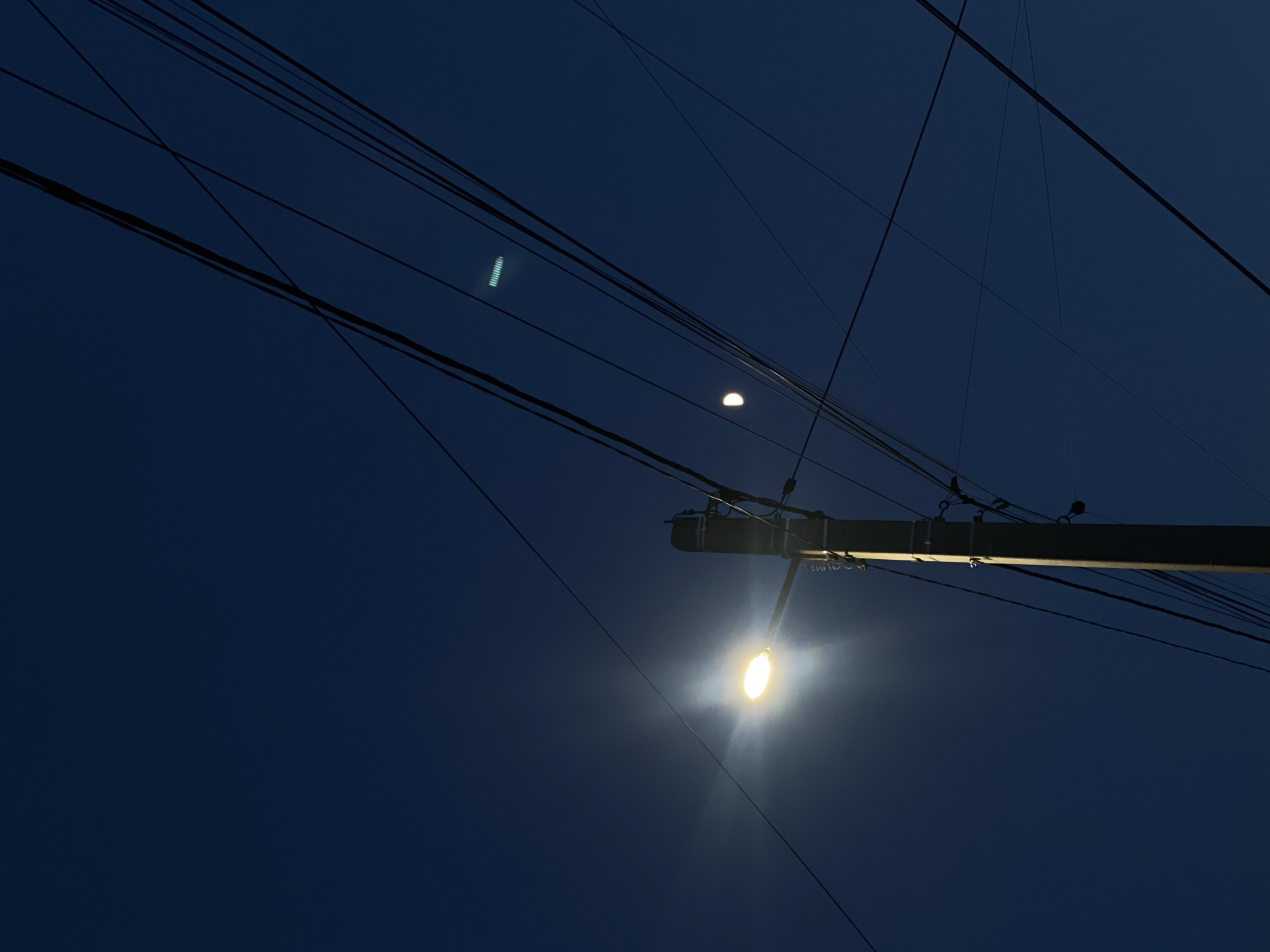 The image captures a dark blue sky at dusk or night, likely in Chișinău, Moldova. Several power lines crisscross the frame, silhouetted against the dim sky. A single street light is prominently featured, illuminating a portion of the scene with a bright, yellow-white glow and creating a starburst effect. The light is attached to a utility pole, which is also visible, along with some of its associated hardware. Another, fainter light source appears higher up and to the left, possibly another distant light or reflection. The overall mood is quiet and somewhat stark, focusing on the mundane infrastructure against the backdrop of the fading daylight. There is no visible text.