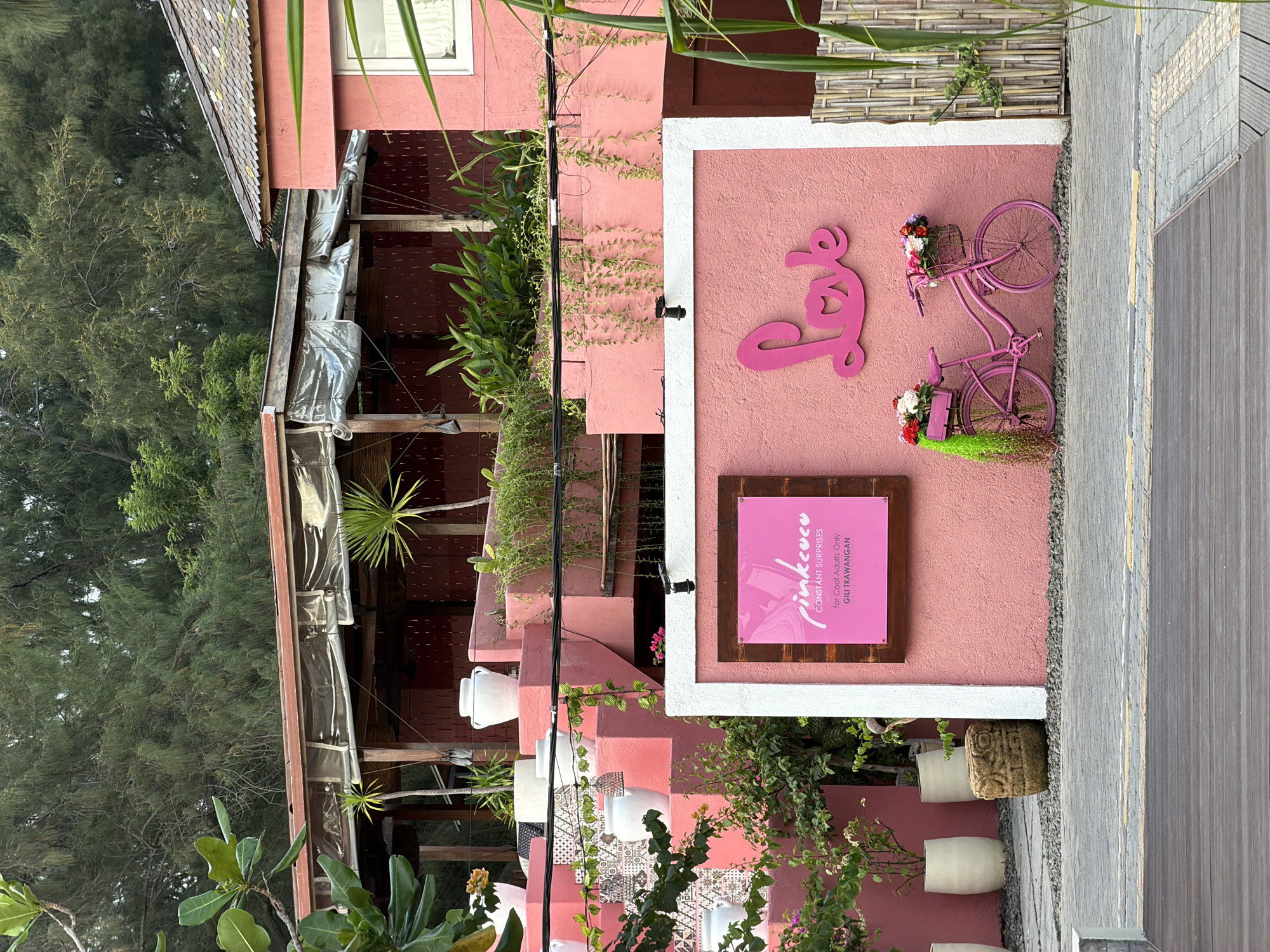 A low-angle, medium outdoor shot captures a pink-themed establishment with an overarching rustic, tropical aesthetic. The building, painted in varying shades of pink, features a multi-level design with white potted plants and vibrant green foliage scattered throughout. In the foreground, a textured pathway leads to a pink stucco wall adorned with a pink wooden sign that reads "pinkcoco CONSTANT SURPRISES for Cool Adults Only GILI TRAWANGAN." To the right of the sign, a pink bicycle is parked, decorated with colorful flowers in its baskets and draped with lush green hanging plants. Above the bicycle, the word "Love" is intricately carved into the wall in a playful, pink cursive font. The background reveals a dense canopy of trees, suggesting a natural, possibly beachfront, environment. The image is devoid of any people or animals.