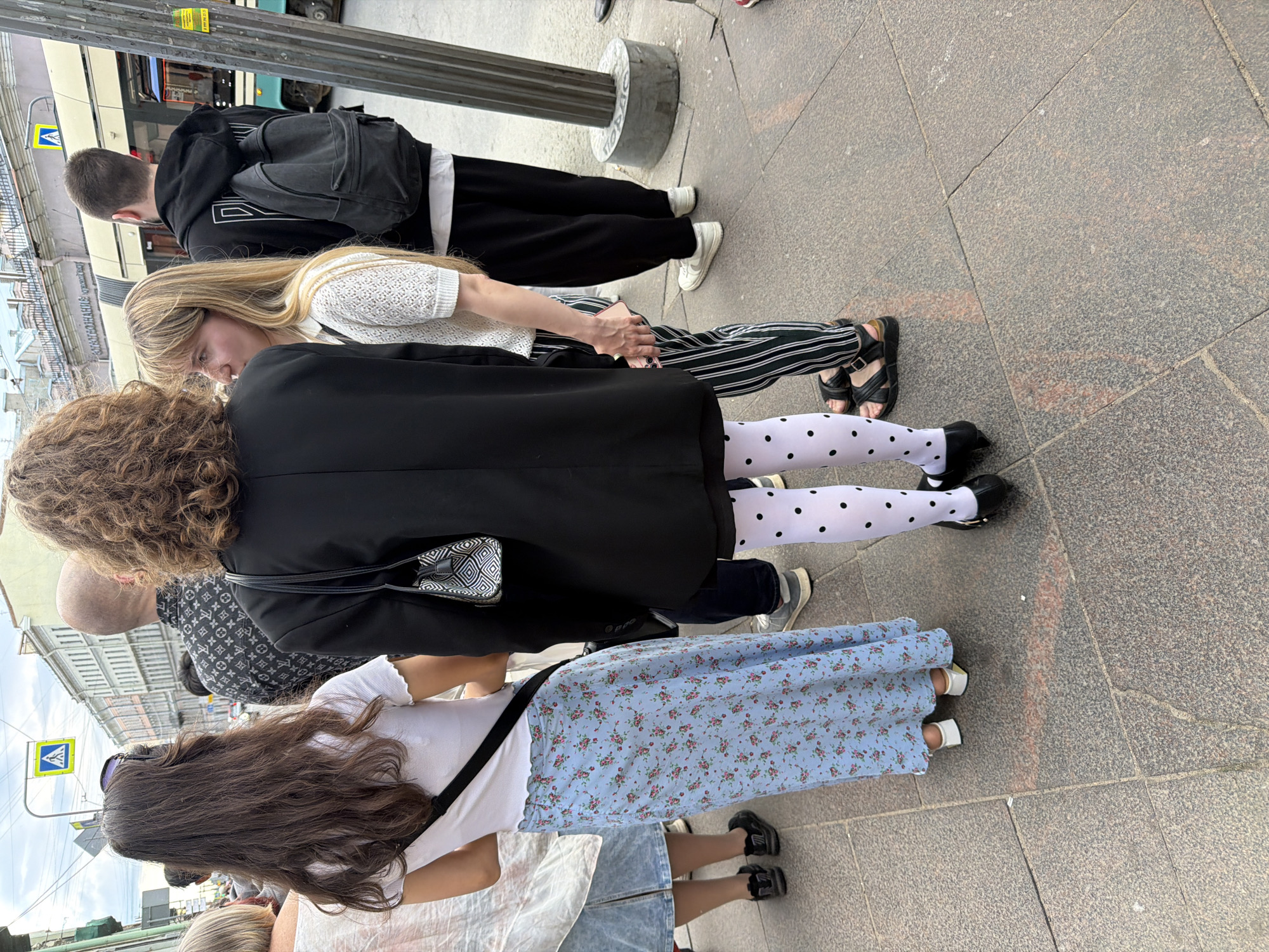 The image depicts a street scene in Saint Petersburg, Russia, likely during daytime given the ambient light. In the foreground, several individuals are visible, primarily from the waist down.
One person, facing away from the camera, wears a black blazer over a white top and a light blue, floral-patterned skirt. They have black and white polka dot tights and black pointed-toe shoes.
To their left, another person, also seen from the back, wears a white t-shirt and a similar light blue floral skirt. They are wearing black sneakers.
To the right of the person in the black blazer, a blonde individual in a white textured top and black and white striped pants stands with their right hand interlocked with the person in the black blazer. They are wearing black sandals. Further to the right, a person in black sweatpants and white sneakers is partially visible.
In the background, buildings with classical architecture line the street. A yellow pedestrian crossing sign is prominently displayed on a pole. A tram or similar public transportation vehicle is visible moving along tracks. The ground is paved with granite slabs. The overall atmosphere suggests a public gathering or a walk through the city.