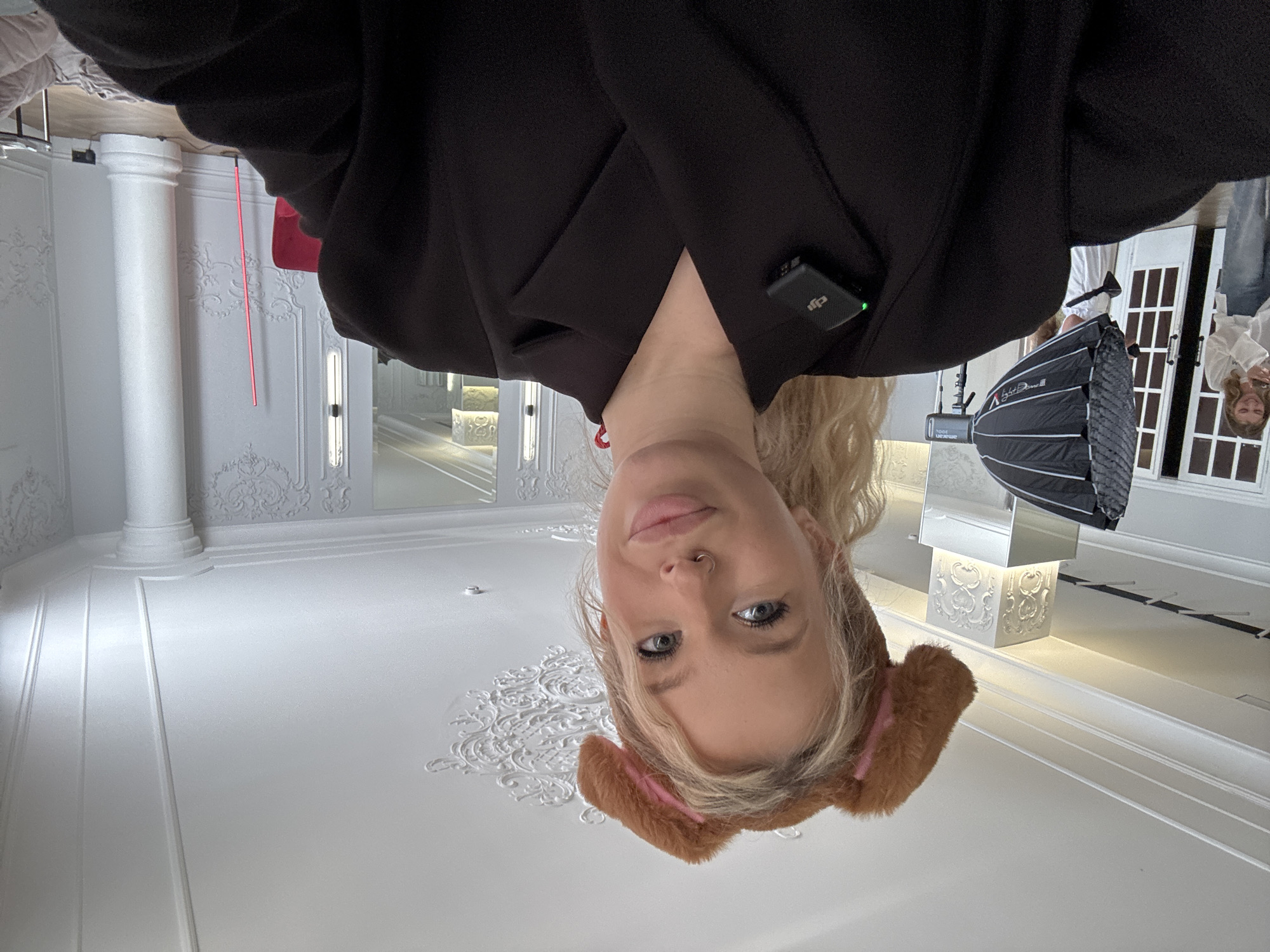This is a low-angle, upside-down selfie of a young woman with blonde hair. She is wearing a black blazer and a fluffy brown headband with pink accents that resemble dog ears. She has a nose ring and light makeup. She is looking directly at the camera with a neutral expression. A small black wireless microphone is clipped to her blazer.
The background appears to be an indoor studio or a room with ornate white moldings and paneling on the walls and ceiling. There are decorative ceiling medallions. To the right, a large softbox lighting setup is visible, suggesting a photo or video shoot is in progress. A white ornate pedestal with a light fixture is also visible. In the distance, there are glimpses of what look like doors with window panes, and reflections in mirrors. The overall impression is a bright, clean, and possibly luxurious setting. The lighting suggests it is daytime or a well-lit interior. There is no visible text in the image.