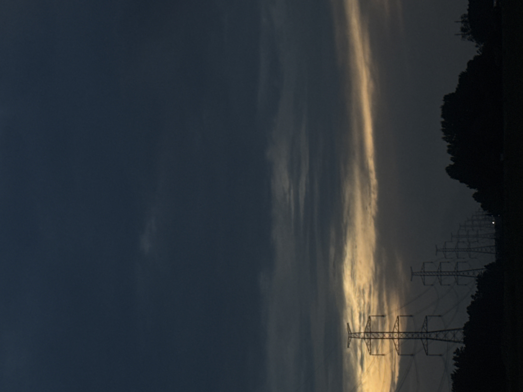 The image captures a twilight scene in Saue, Estonia. A dark, predominantly blue-gray sky, heavily overcast with stratus clouds, occupies the upper two-thirds of the frame. A horizontal band of diffuse yellow-orange light is visible on the horizon, suggesting either sunrise or sunset. Below this lighter band, a series of large, metallic electricity pylons (transmission towers) are silhouetted against the sky, extending from the lower-left to the mid-right in a receding perspective. The closest pylon, positioned in the lower-left, is the largest and most detailed, exhibiting its structural framework and power lines. Subsequent pylons diminish in size and clarity towards the right. Below the pylons, a dark, undulating silhouette of trees and dense vegetation forms the immediate horizon, obscuring any ground details. The foreground is dark and indistinct. No people, animals, or vehicles are visible.