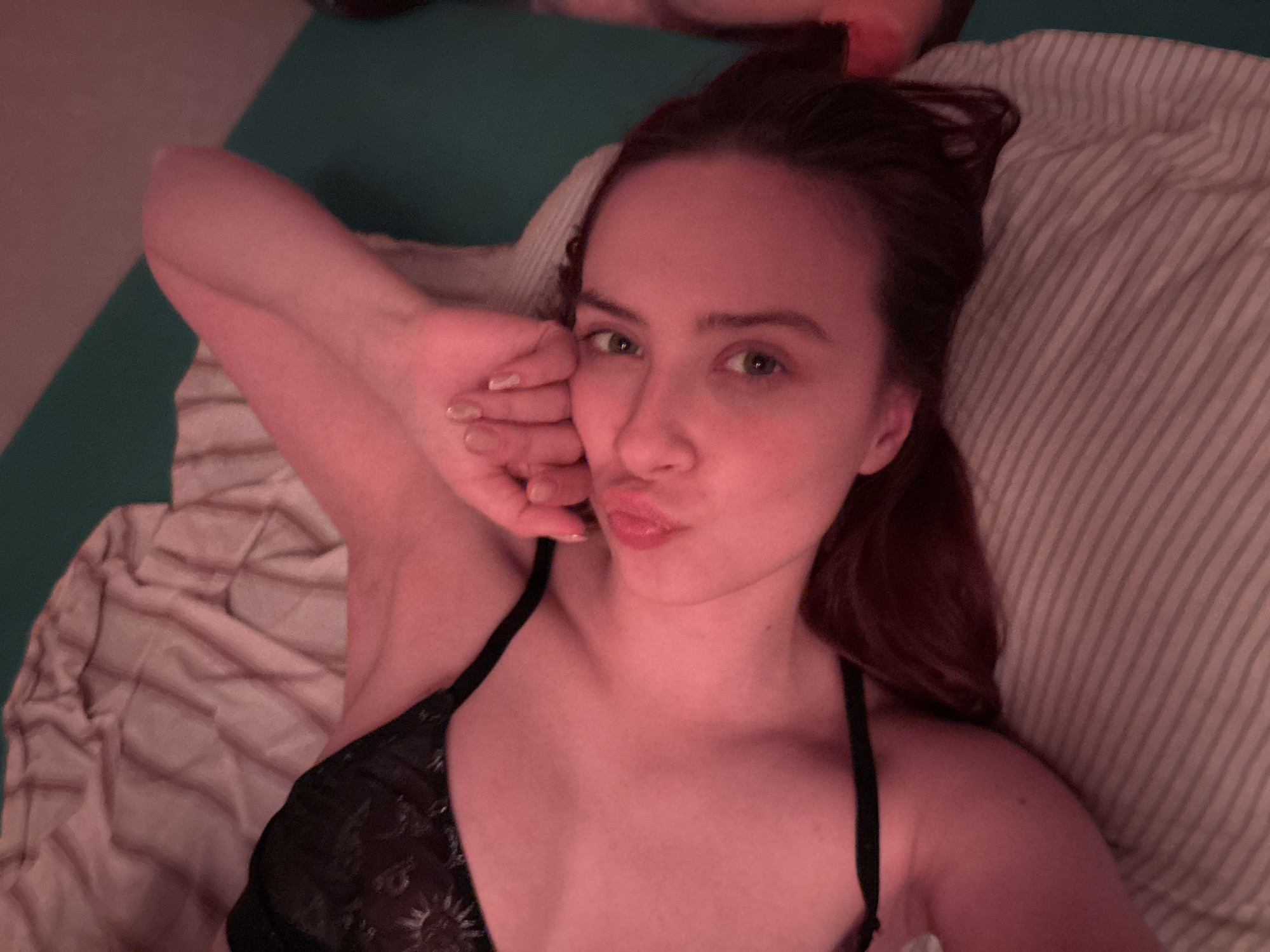 A young woman with fair skin, green eyes, and dark hair is depicted lying on a bed, appearing to be indoors. She is captured in a close-up, top-down perspective, looking directly at the camera with puckered lips in a playful expression. Her left arm is bent, with her hand resting near her face, revealing light-colored nail polish. She is wearing a black top, possibly a bra or camisole, featuring an intricate, possibly floral or starburst-like, lace or embroidered pattern.
Her head rests on a pillow with prominent light-colored stripes, while a plain green sheet or duvet is visible behind her head and to the upper left. Another lighter, textured sheet is present towards the bottom left of the frame. The entire scene is illuminated by a warm, pinkish-red light, casting a distinct hue over her skin and the surroundings, suggesting artificial lighting or a color filter, likely indicating an evening or nighttime setting. There is no visible text in the image. The implied location is Прикамский, Russia.