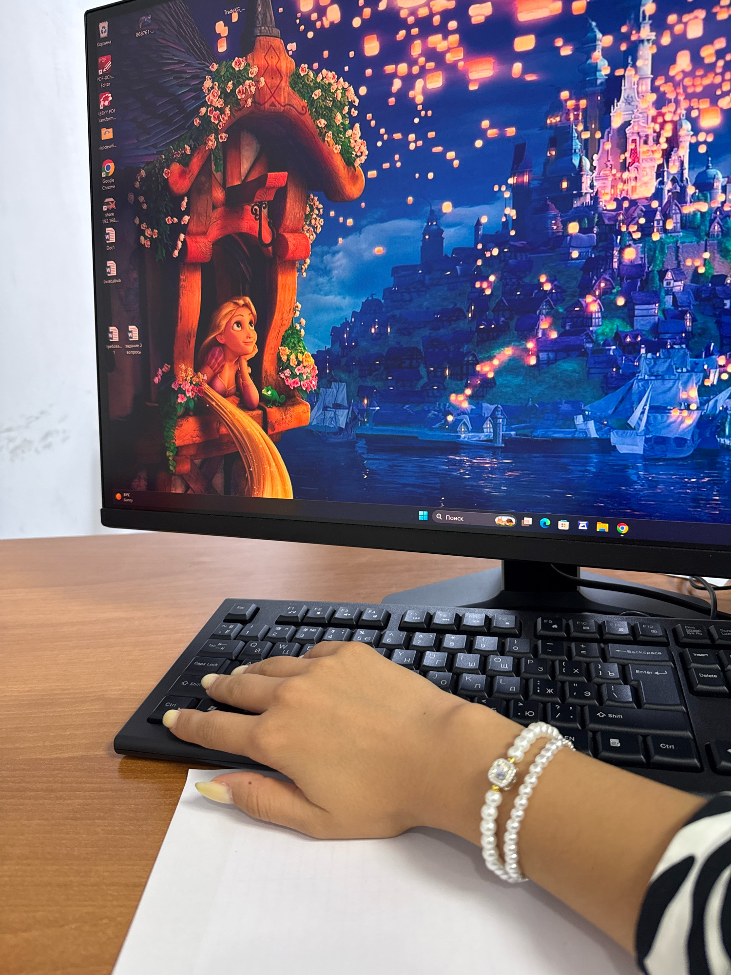 A computer monitor displays a scene from the movie Tangled, featuring the character Rapunzel and a cityscape at night illuminated by lanterns. In the foreground, a person's hands rest on a black computer keyboard, wearing a pearl bracelet. The scene is set on a wooden desk, suggesting an indoor office or home environment. There is no visual information to determine its location in Bishkek City, Kyrgyzstan.