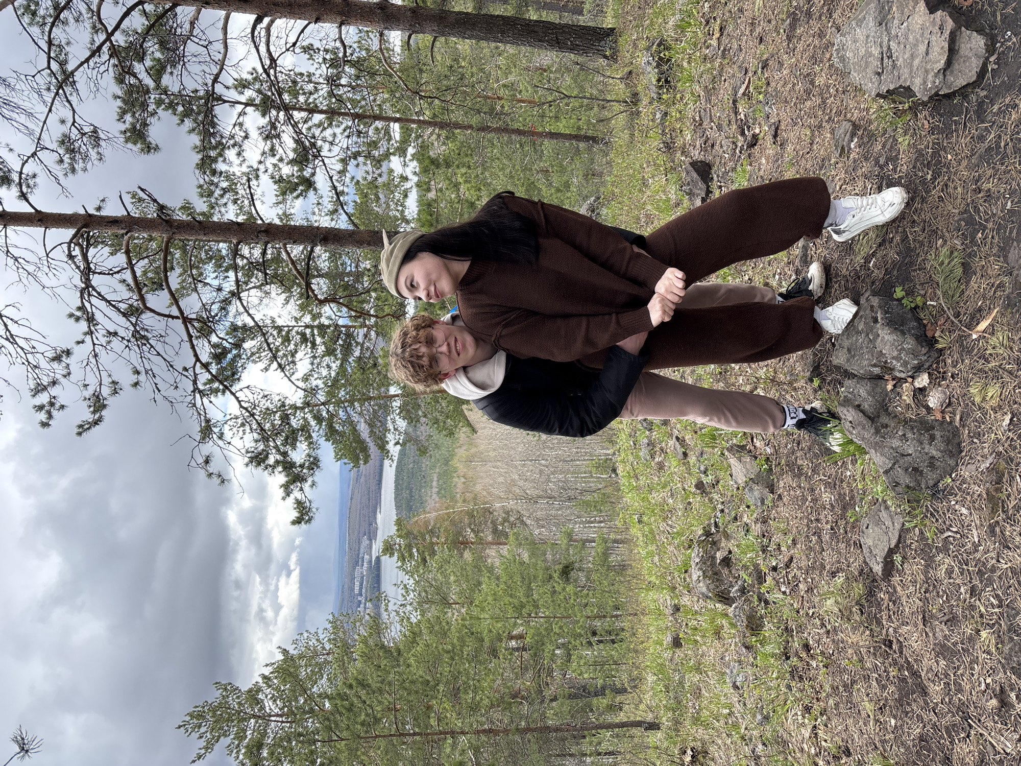 Two people are embracing outdoors. One person is wearing a brown sweater and pants, and a green cap. The other person is wearing a black jacket and light brown pants. They are standing on a hillside amongst trees and rocks. A body of water and city buildings are visible in the background. The location is in or near Kachkanar, Russia.