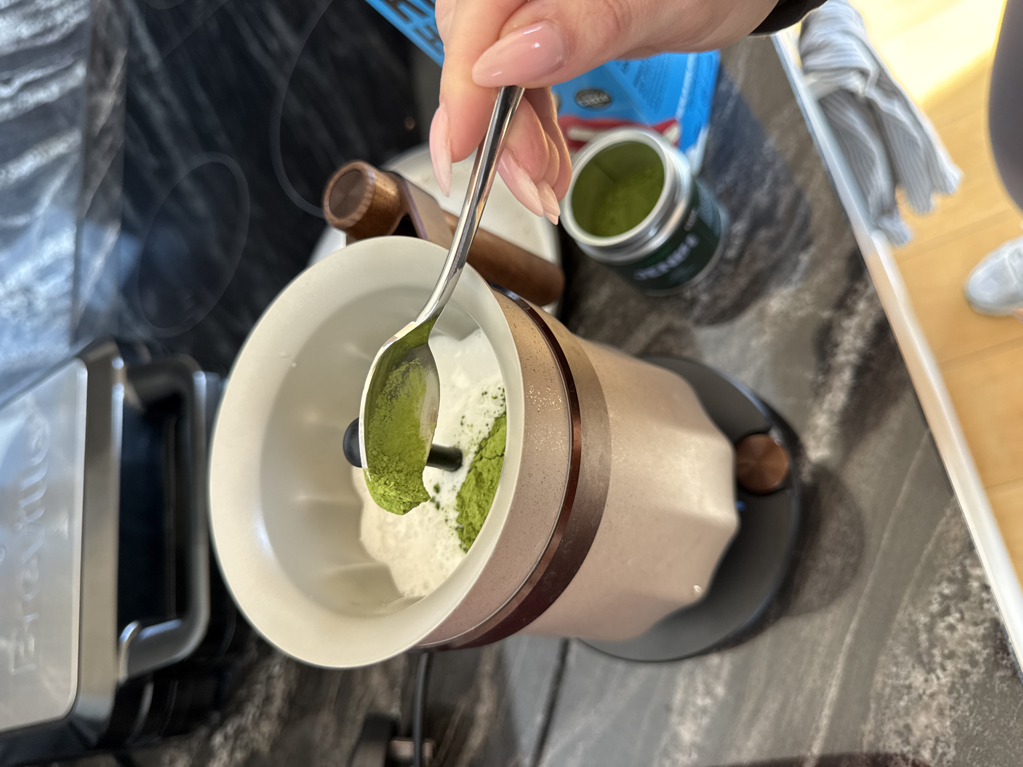 In a kitchen located in the city of London, United Kingdom, a person's hand, displaying light pink manicured nails, holds a silver spoon, in the act of depositing green matcha powder into a cream and bronze-colored Breville milk frother that contains a white, foamy liquid. A small quantity of green matcha powder is already visible at the bottom of the frother's contents. The frother is positioned on a dark, mottled countertop, adjacent to a black induction hob featuring multiple circular elements. In the background, a distinct Breville appliance, appearing to be a grill or panini press, is partially visible. Also present on the countertop are an open tin of green matcha powder with a silver rim, a small wooden-handled tool, a blue packaged item, and a striped cloth. The lower right foreground shows a portion of a foot clad in a light-colored shoe.