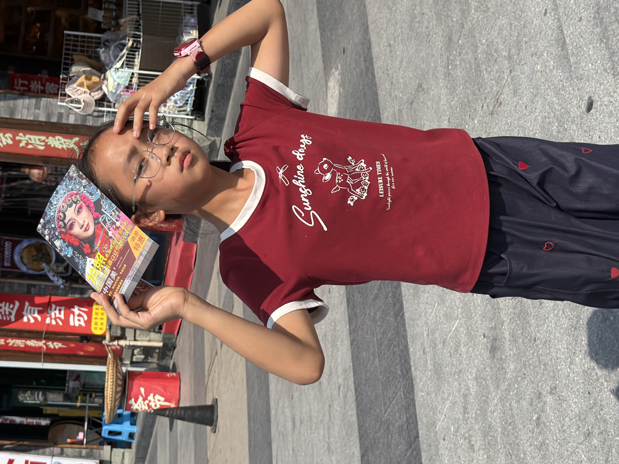 A young girl wearing glasses and a red t-shirt with a deer graphic and text that reads "Sunshine days LEISURE TIMES Sunlight pierces through the world of treasured fliers over summer" stands outdoors, holding a book over her forehead to shield herself from the sun. She is wearing dark blue pants with a red heart pattern. In the background, there are shops with red banners, and a street or plaza paved with grey stones. The overall scene suggests a commercial area in the city of Shapingba District, China, likely during the daytime due to the bright sunlight.