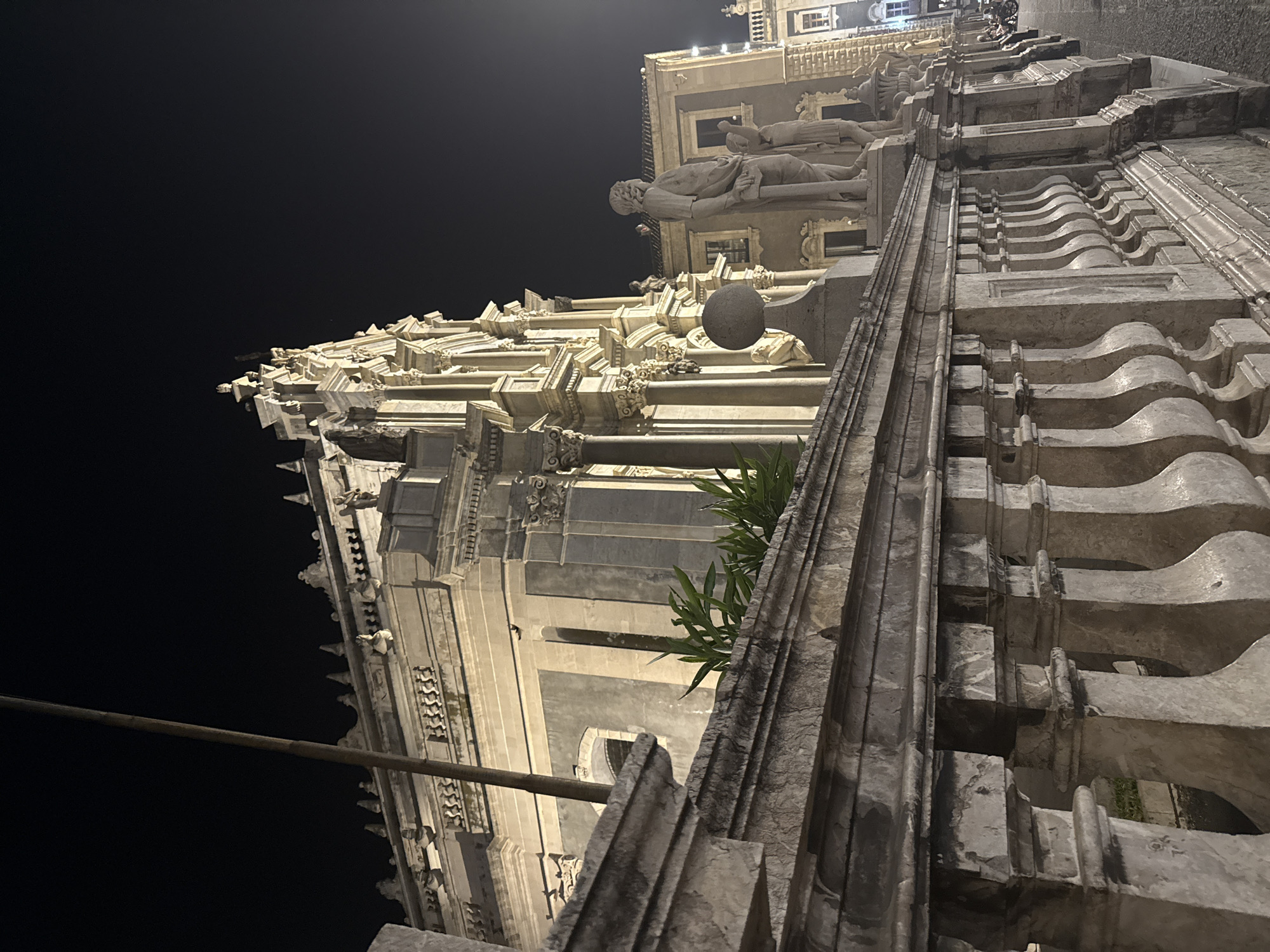Captured at night, the image showcases a building with intricate architectural details, possibly a church or significant historical structure. The building's exterior is adorned with statues, carvings, and ornate stonework, illuminated by artificial lighting. The foreground is dominated by a close-up view of architectural elements, such as balustrades and columns, constructed from stone. Above, the middle ground features a statue atop the building and a series of illuminated windows. In the upper portion of the image, the night sky provides a dark contrast to the light-colored building.