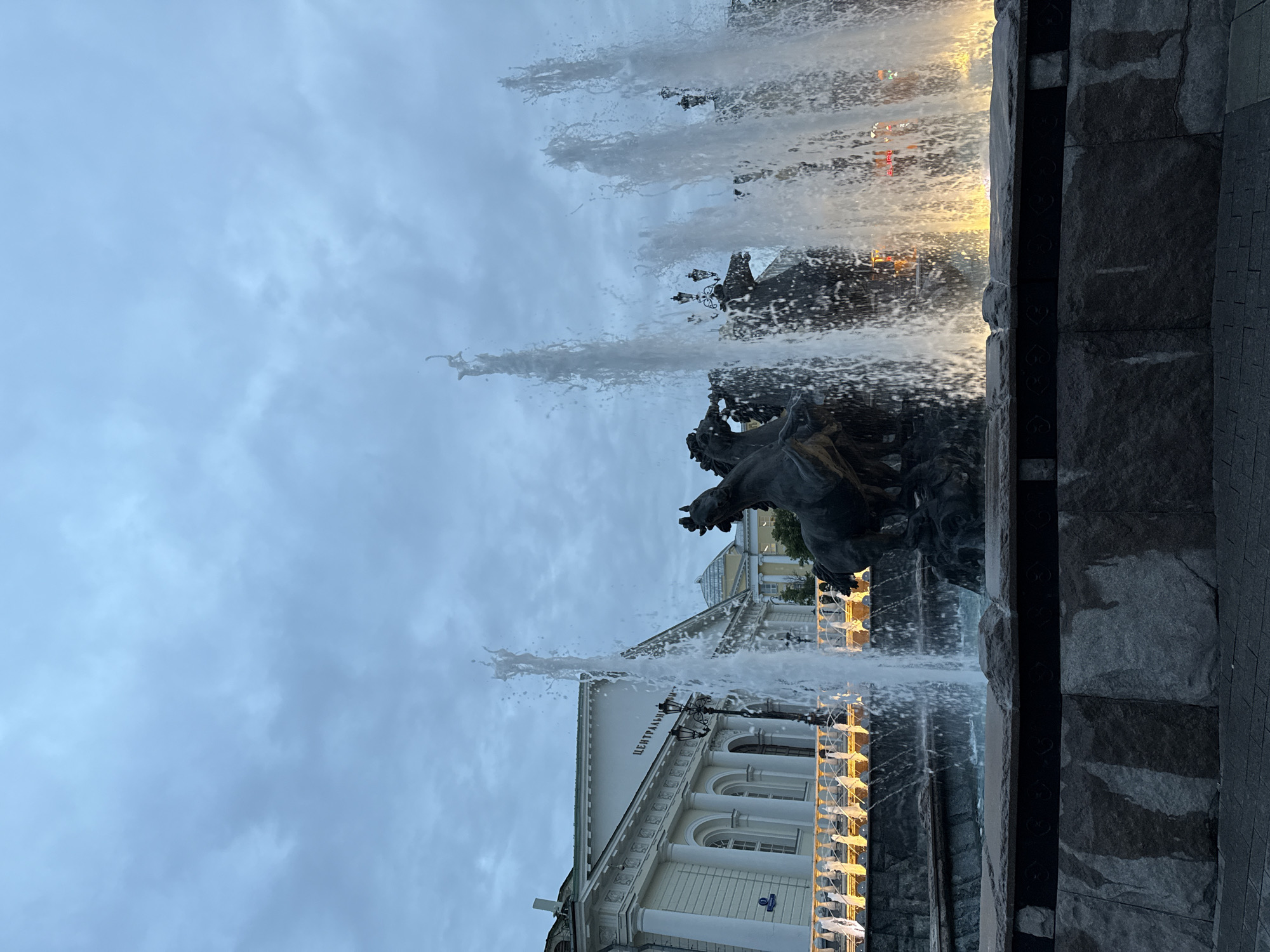 The image captures a fountain in Moscow, Russia, featuring a sculpture of a horse-drawn chariot. Water jets upward, partially obscuring the view of a building behind the fountain, identified by lettering (partially visible as "ЦЕНТРАЛЬНЫЙ") as the Central Children's Music Theater. The sky is overcast. The photograph appears to be taken from a low angle near the base of the fountain. The wall of the fountain is made from dark stone. Lighting illuminates the fountain and the building's architectural details.